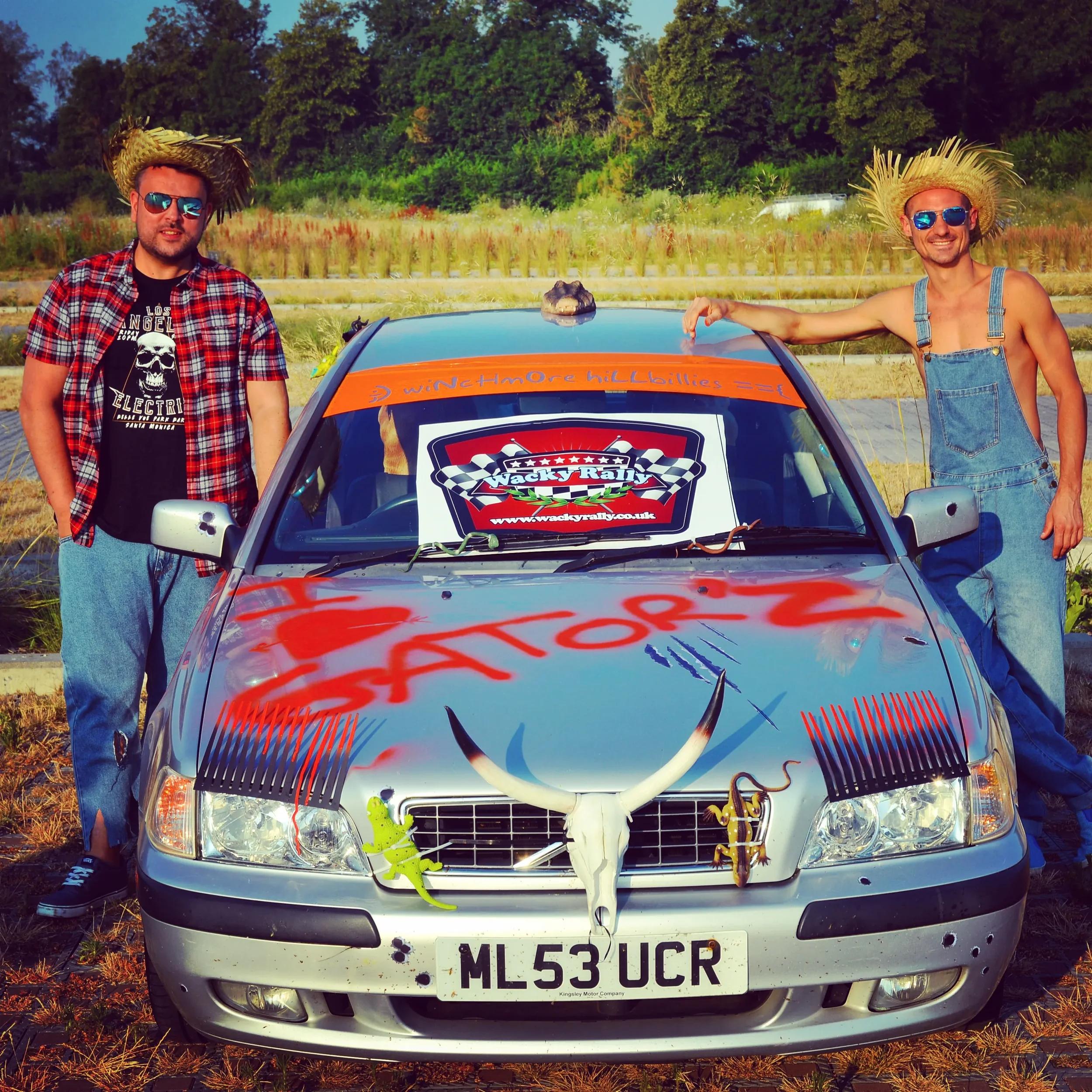 Two men wearing straw hats and sunglasses stand beside a decorated silver car. The car's hood and front are spray-painted with red graffiti. A sign on the windshield reads 'Wacky Rally'. The front of the car has various decorations, including a large