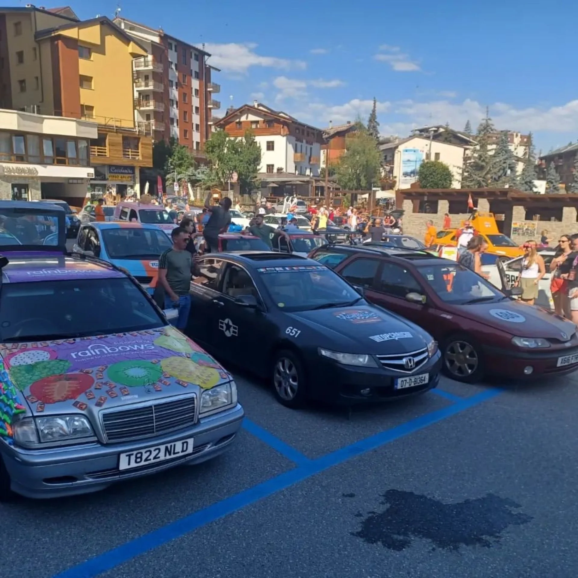A parking lot filled with various cars, surrounded by people and buildings on a sunny day.