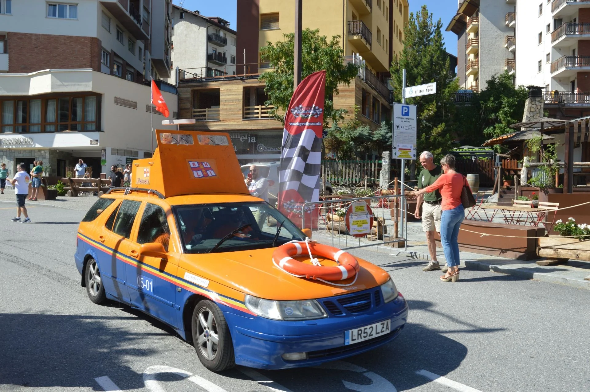 Orange and blue emergency or race car with a life preserver on its hood, parked on a city street with people walking and talking nearby, modern buildings surround the scene.
