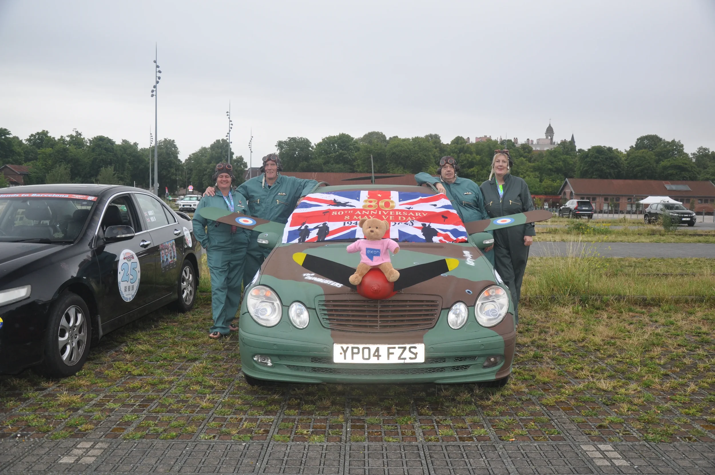 Four women in vintage pilot costumes standing behind a decorated military style car with a teddy bear on the hood, during a commemorative event with other cars and flags in the background.
