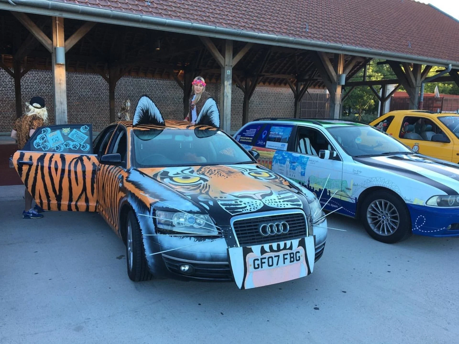 A black Audi car with tiger face artwork on the hood and tiger stripes on the body, featuring a large tiger face on the front of the car and tiger ears on the roof, parked in a lot alongside another decorated car with a scenic wrap.