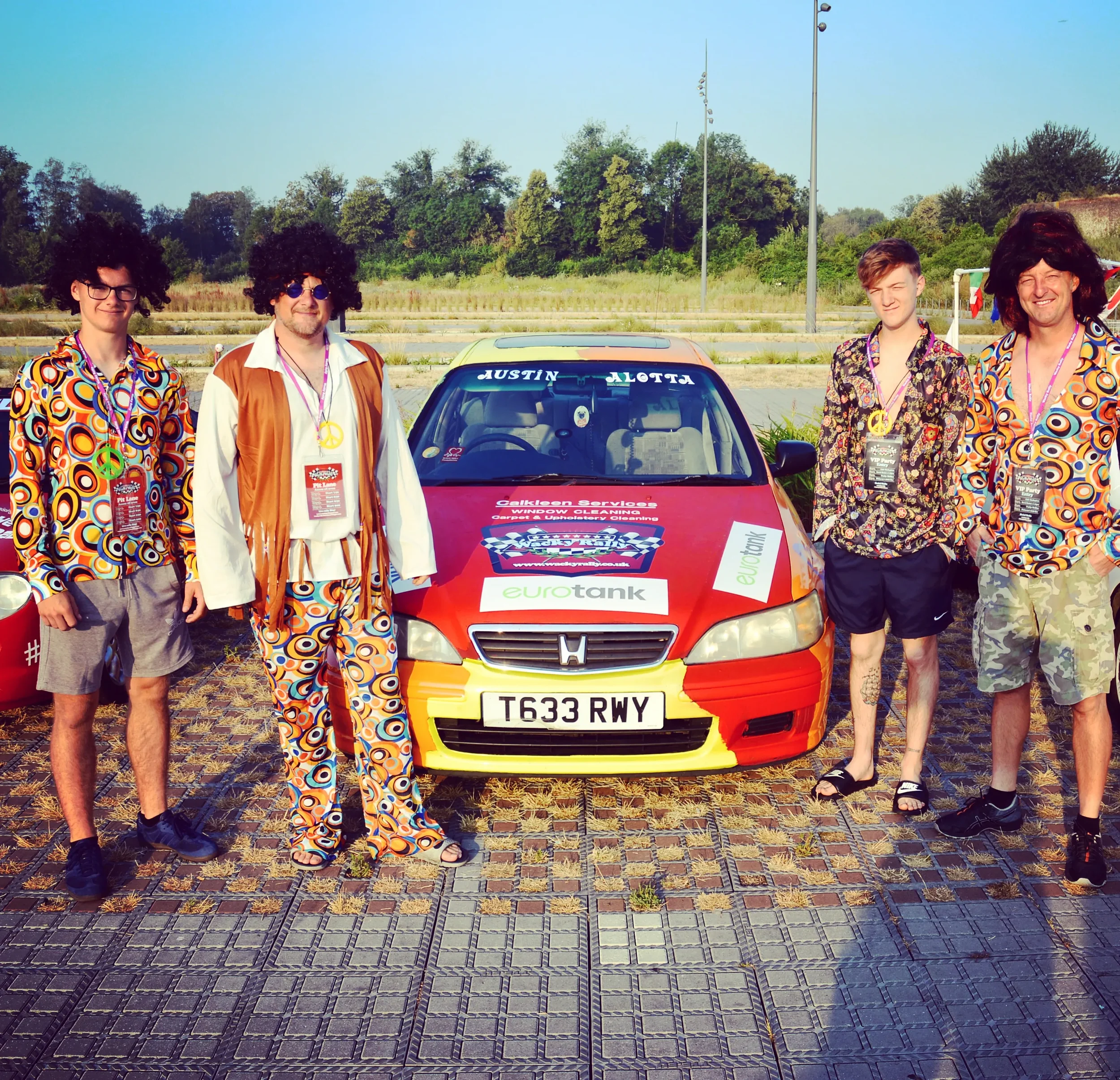 Five people dressed in colorful 1960s-style clothing and wigs standing beside a small red car with racing and sponsorship decals, on a paved outdoor area with trees and streetlights in the background.