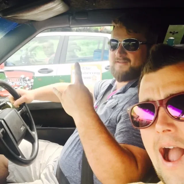 Two men in a vehicle, one with sunglasses and a beard, pointing backward with his hand, the other with sunglasses and an open mouth, both smiling, with a parking lot and other vehicles visible outside the window.