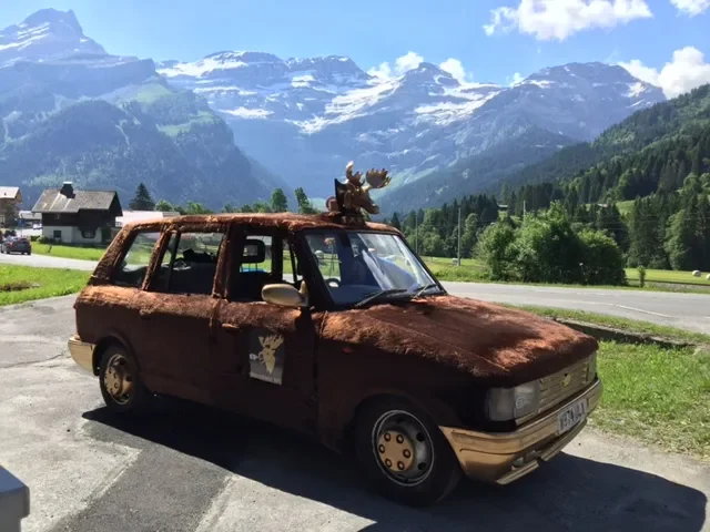 Rusty, weathered car with a reindeer figure on top, parked near a scenic mountain landscape with snow-capped peaks and green hills.