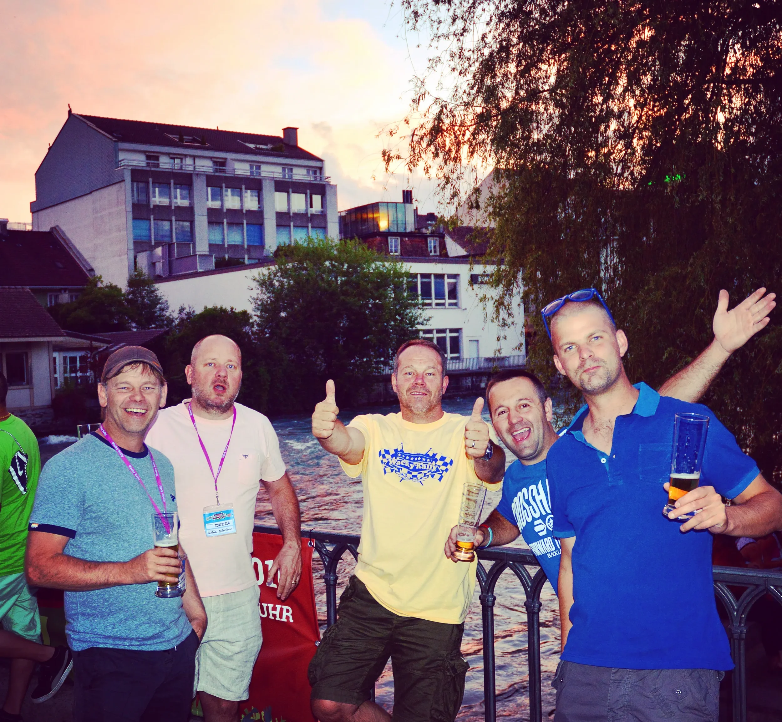 Group of five men enjoying drinks outside near a river at sunset, smiling and posing for the photo.