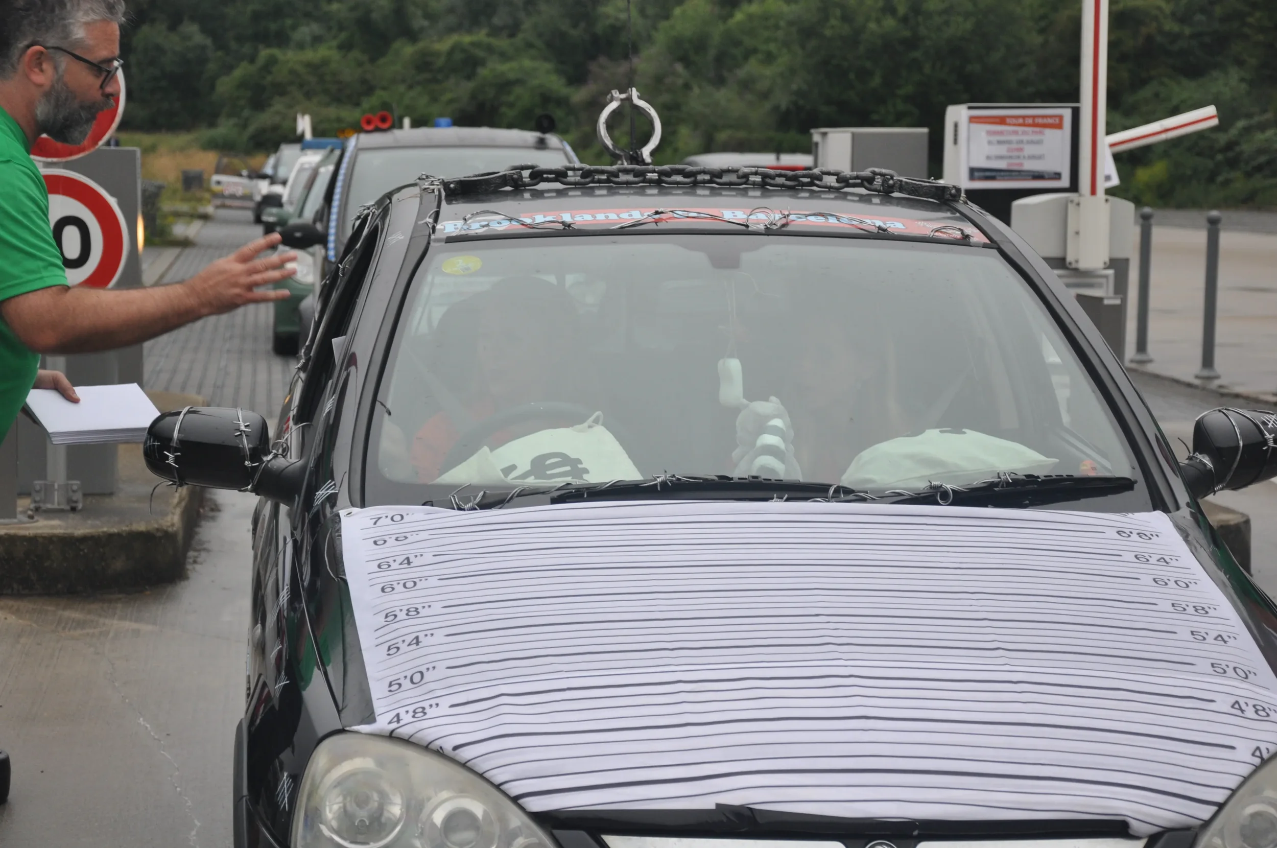 A man with glasses and a green shirt is talking to a car at a toll booth. The car has a height measurement chart covering its hood, and there is a glass partition with a figure giving a thumbs-up inside the vehicle.