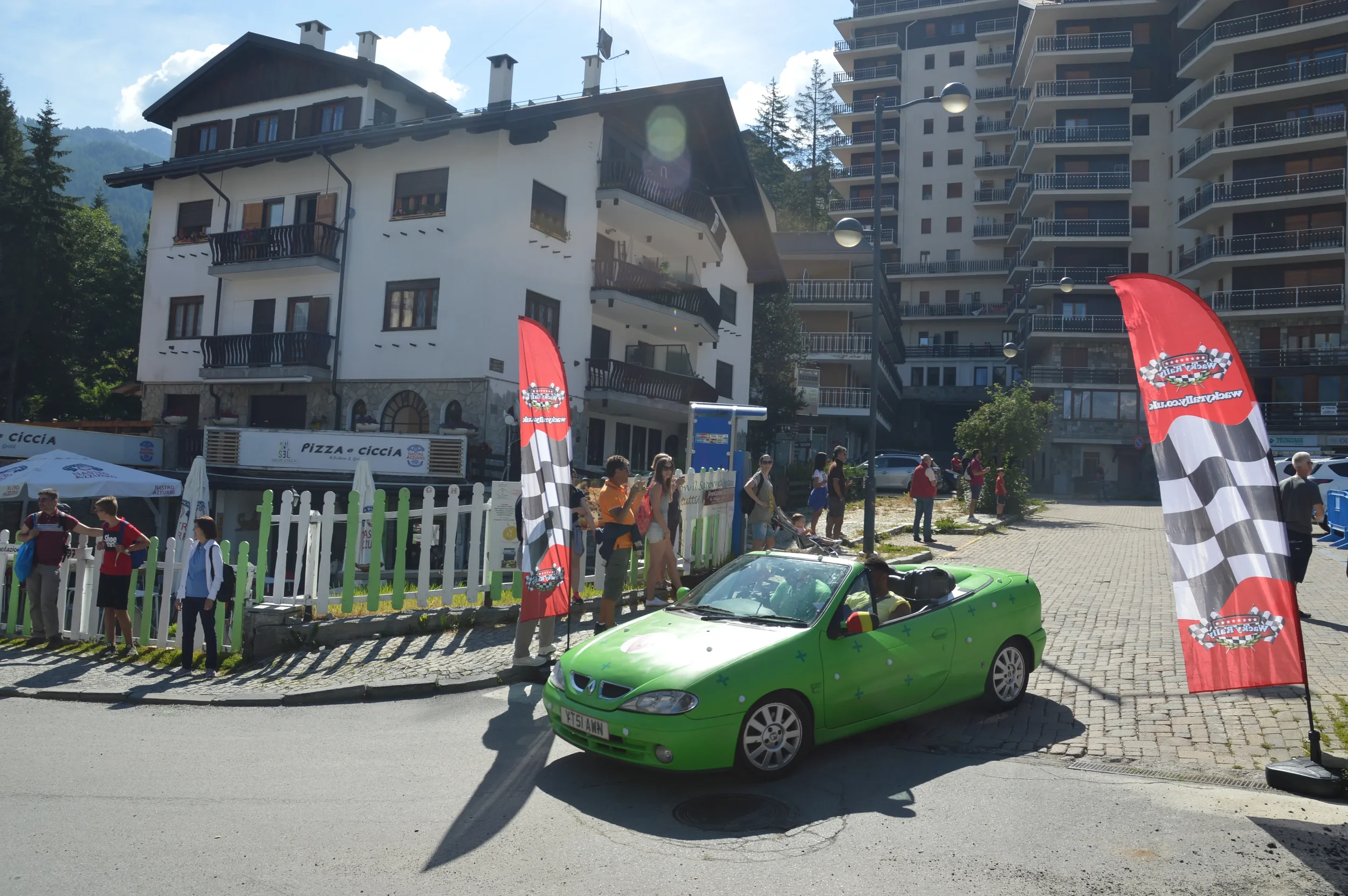 A bright green convertible car with a driver inside, parked on the street near a group of people gathered around. Two red flags with checkered patterns are positioned on either side of the scene. Behind, there are several multi-story buildings, inclu
