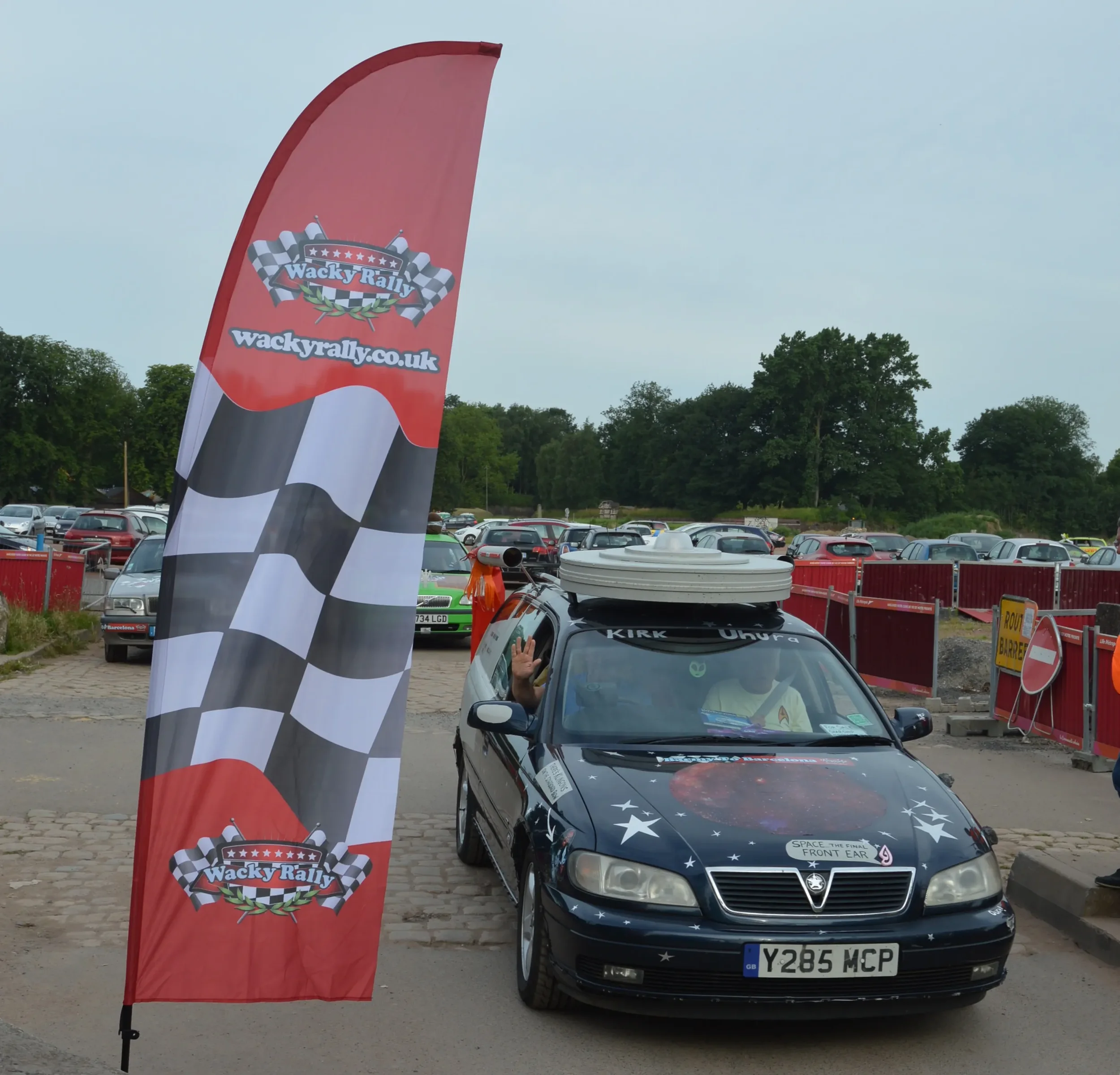 A black car with space-themed decorations and a front bumper sticker that says 'SPACE THE FINAL FRONT EAR' parked next to a red and black checkered flag banner that reads 'Wacky Rally.' The car driver is waving from the window, and the car has a larg