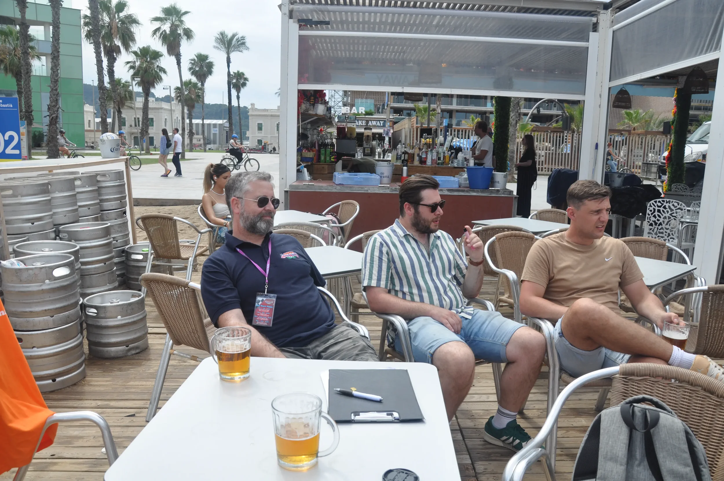 Three men sitting outdoors at a bar, each with a glass of beer. Two are wearing sunglasses, one in a tan Nike T-shirt, another in a striped shirt, and the third in a navy polo. Behind them, a woman is sitting at a nearby table. There are stacked beer
