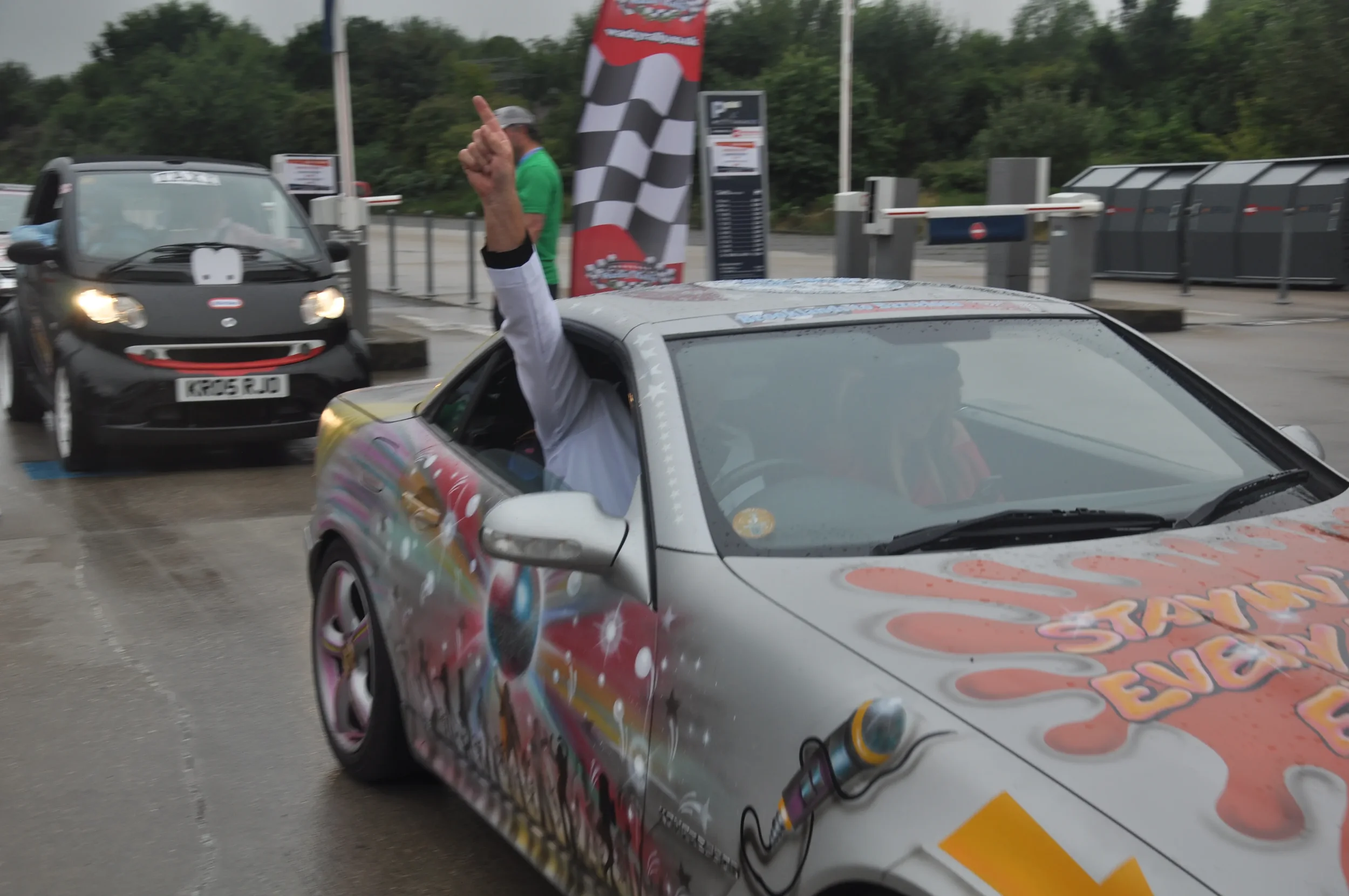 Person in a spray-painted car giving a thumbs-up and raising a hand with a finger pointing up at a race track