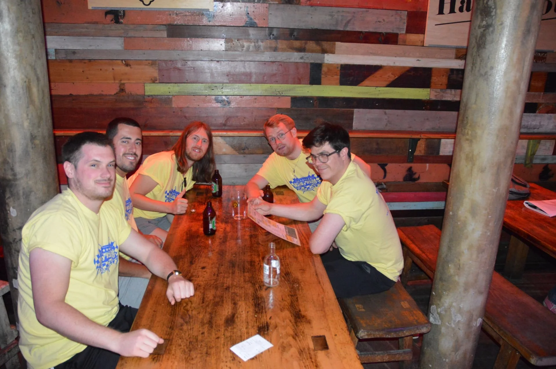 Five men sitting at a wooden table inside a rustic bar or restaurant, wearing matching yellow shirts, with bottles and drinks on the table.