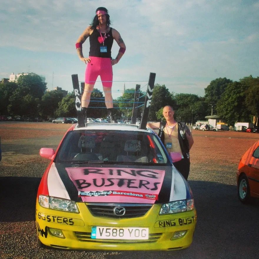 Person dressed as a wrestler standing on the roof of a decorated car labeled 'Ring Busters,' parked in an open area with trees and other cars in the background.