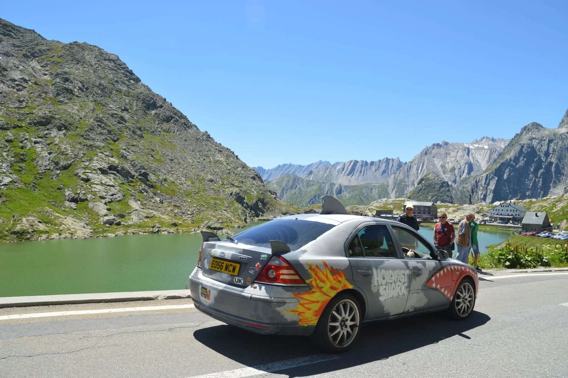 A silver car with fantasy dragon-themed paintwork, including the words 'HOBBf SHARK,' parked on a mountain road near a green lake, with a group of people standing nearby and mountains in the background under a blue sky.