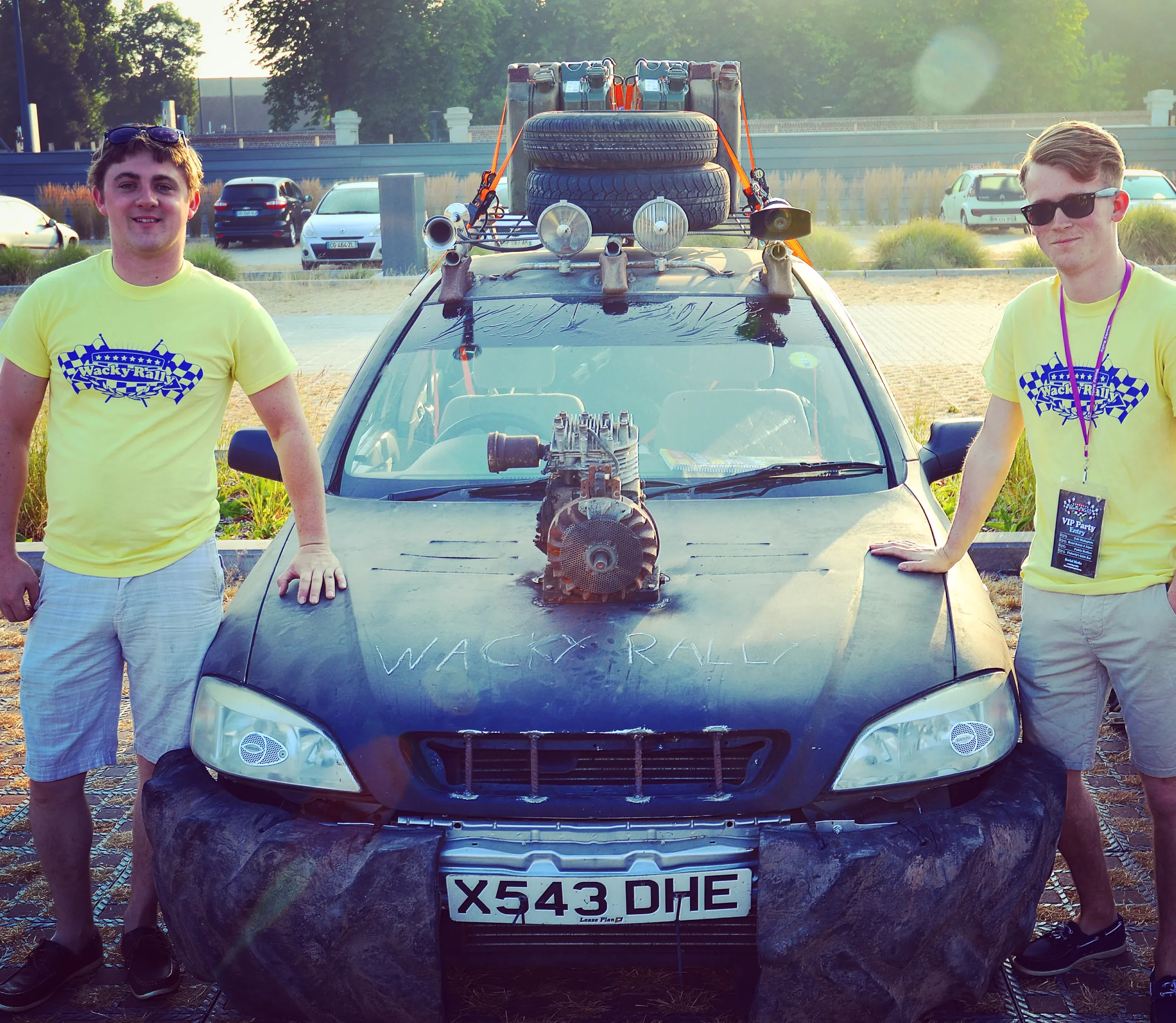 Two young men standing beside a modified car with a large engine on the hood, surrounded by a black tarp, at an outdoor event during daylight.