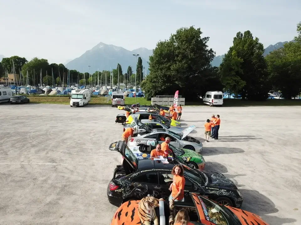 A row of sports cars with open hoods and trunk lids parked on a gravel lot near a marina with sailboats and a mountain backdrop. Several people in orange shirts are inspecting and discussing the cars.