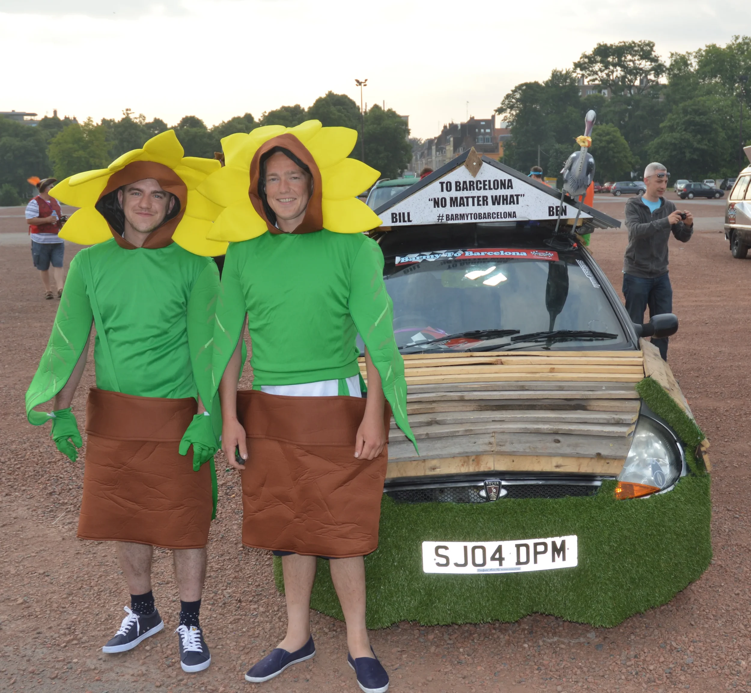 Two people dressed as sunflowers stand in front of a decorated car with a sign that says 'To Barcelona 'No Matter What''. The car is decorated with wooden planks, artificial grass, and a plastic stork figure. Several other people are visible in the b