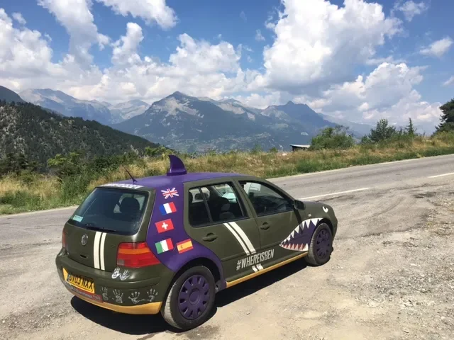 A car decorated with shark teeth graphic on the side and stickers of flags, parked on a gravel side of a mountain road with scenic mountains and sky in the background.