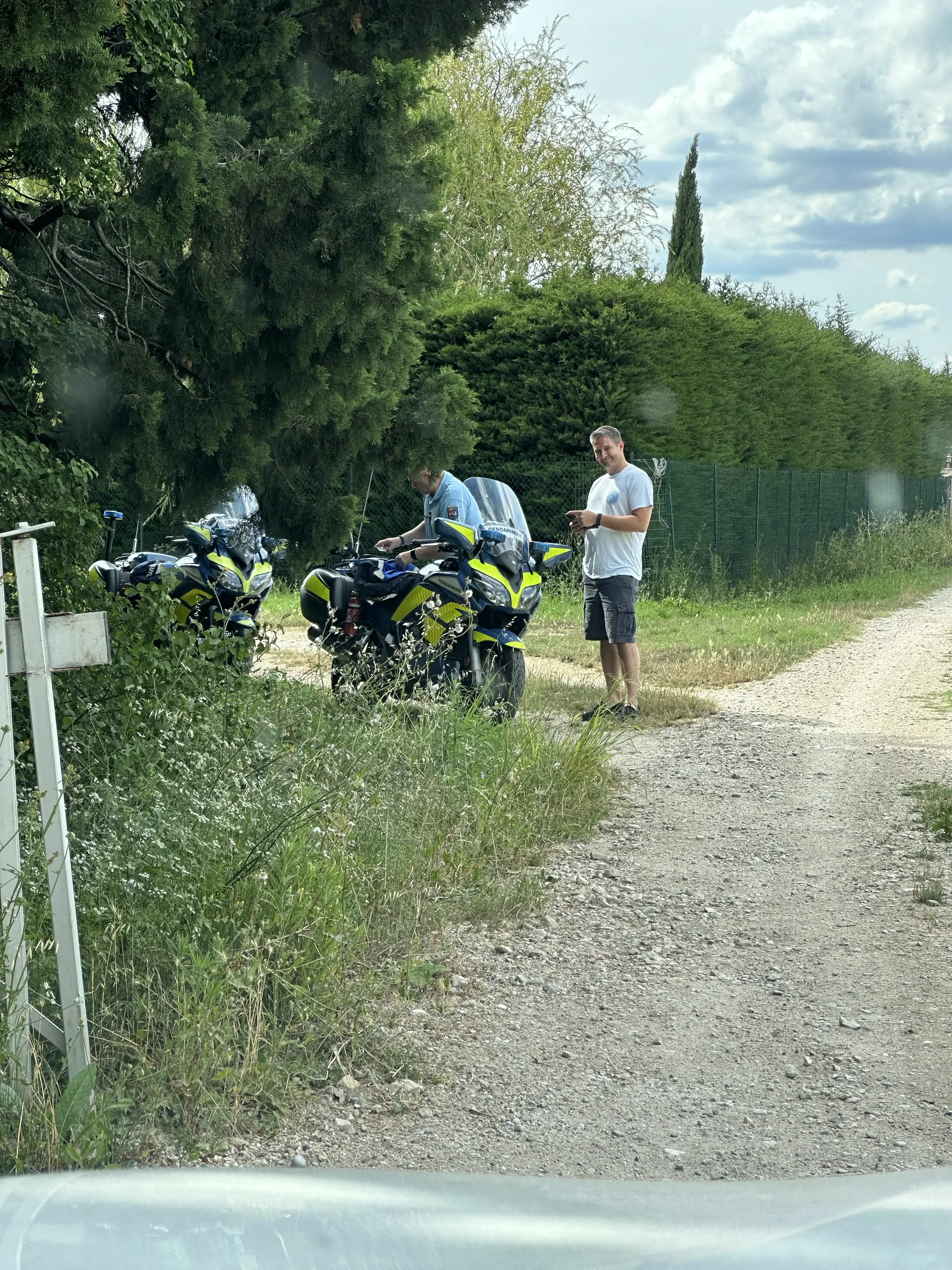 Two police officers checking motorbikes on a rural roadside while a man, dressed in a white t-shirt and shorts, looks on and holds a mobile phone. The scene is set under a cloudy sky with green trees and bushes in the background.