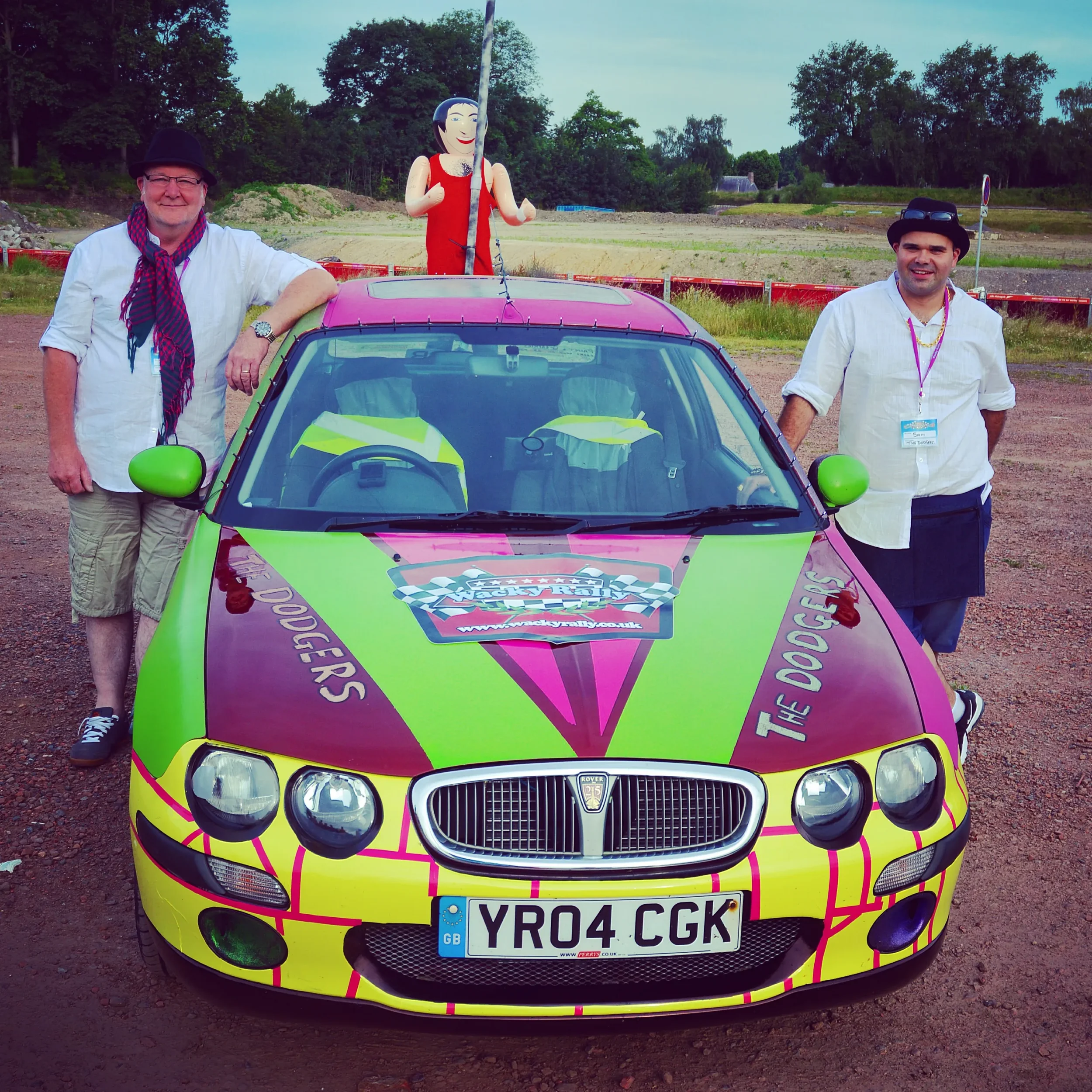 Two men standing next to a colorful yellow, pink, and green Dodge car with a NASCAR logo on the hood. One man is on the left, wearing a black hat, glasses, white shirt, shorts, and a scarf. The other man is on the right, wearing a black hat, white sh