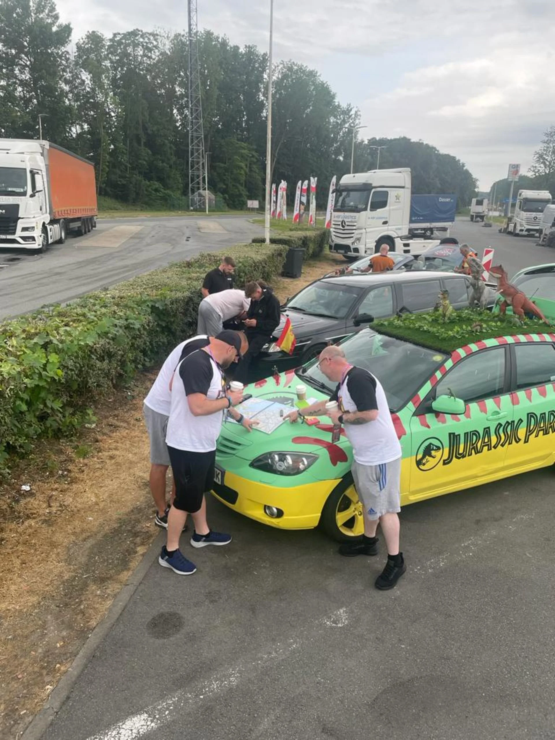 People gathered around a 'Jurassic Park'-themed car with dinosaur decorations, signs of a meetup or event, near a highway with trucks in the background.