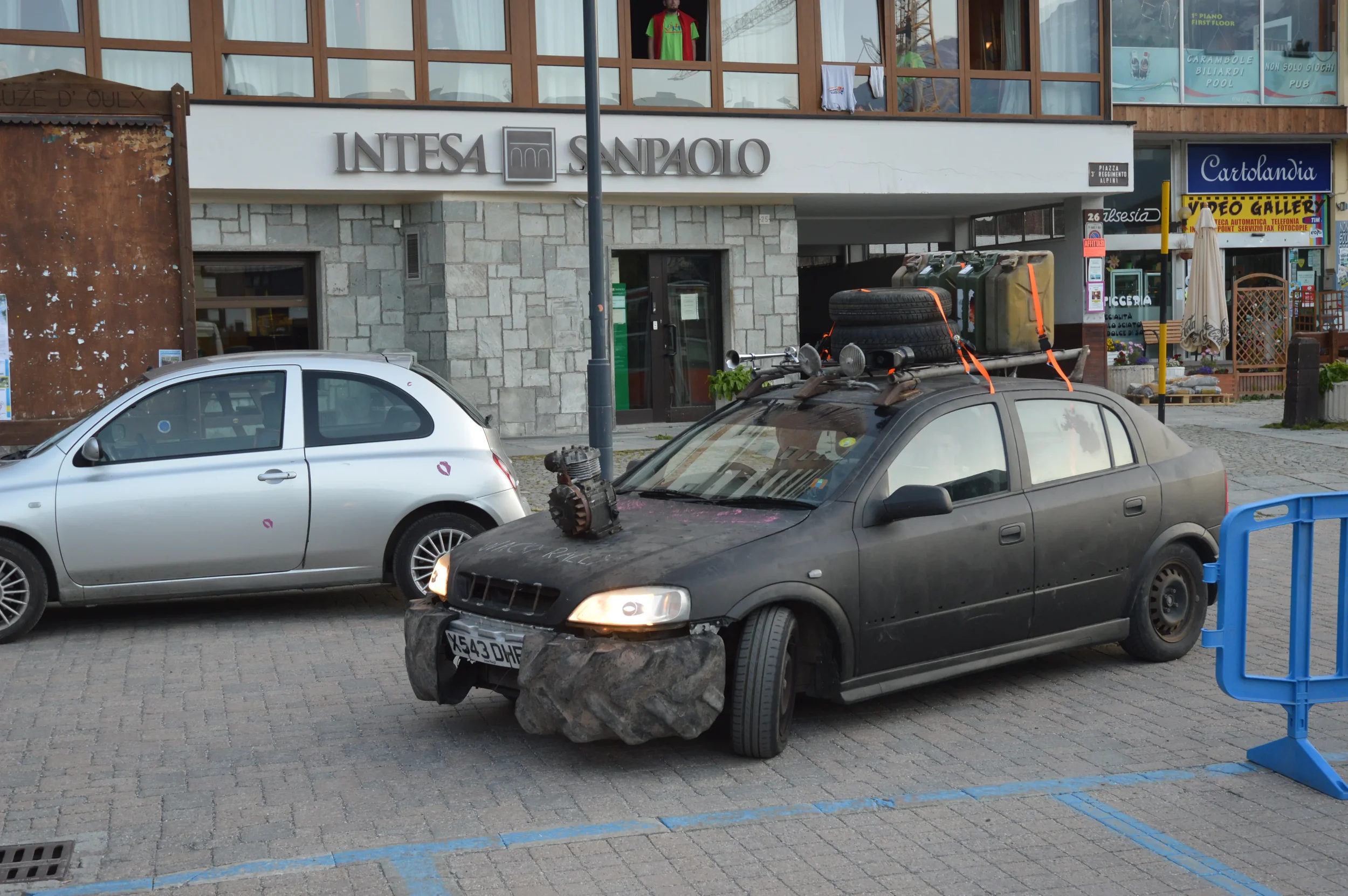 A black car with a large rock attached to the front bumper, with additional objects and luggage on its roof, parked on a cobblestone street beside a silver car, in front of a building with a sign that reads 'INTESA SANPAOLO'.