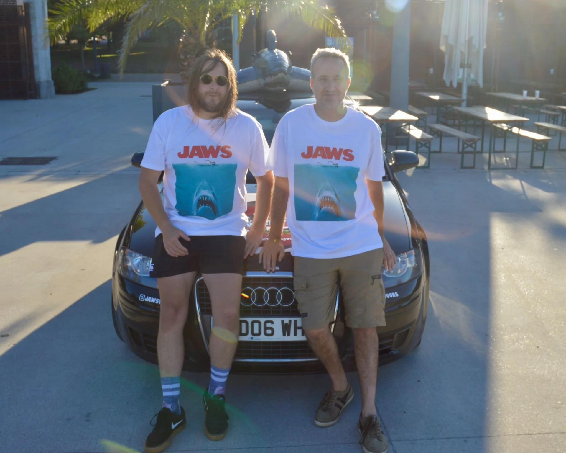 Two men standing in front of a black Audi car, both wearing white 'Jaws' T-shirts with shark image, casually dressed, outdoors in a sunny area with tables and a statue of a turtle in the background.