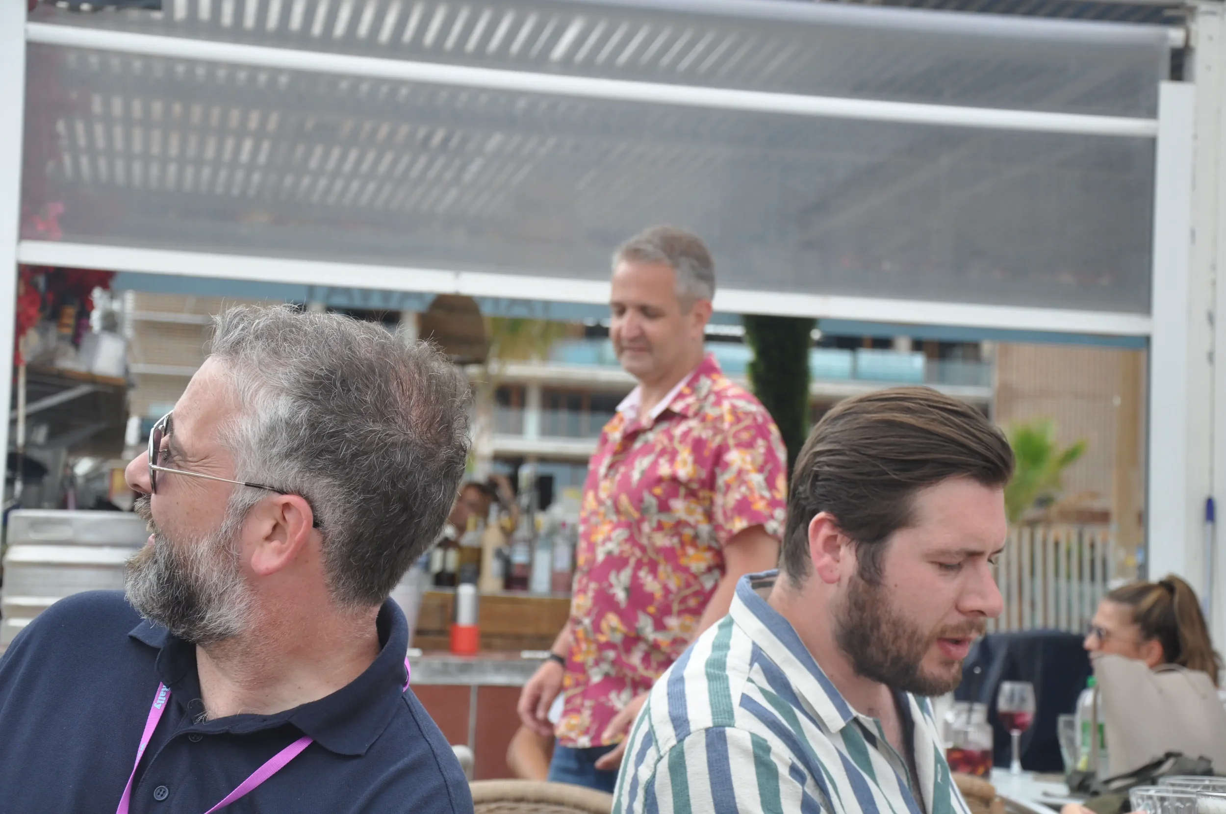 People socializing at an outdoor dining area, with one man in a tropical shirt standing in the background.