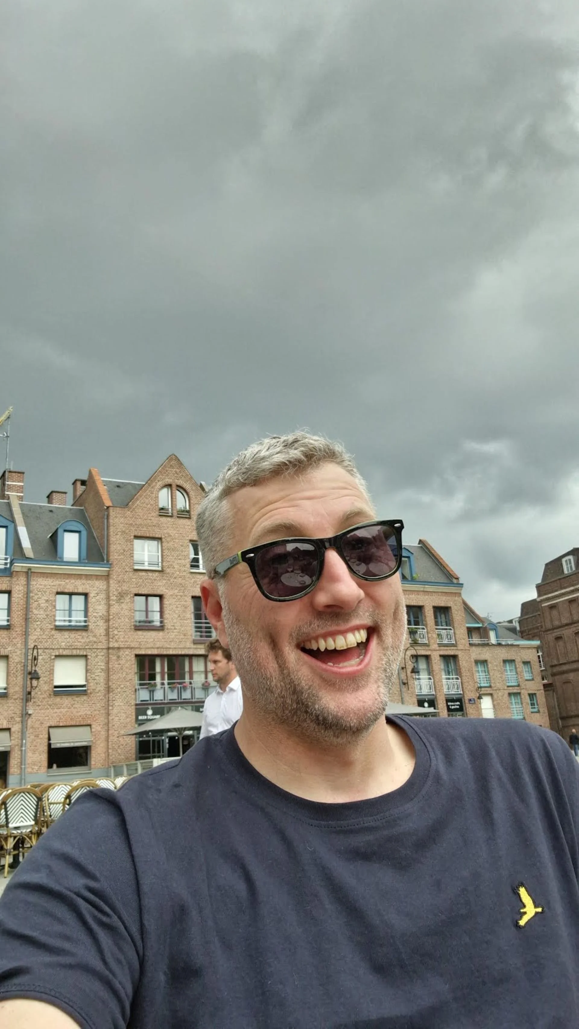 A man smiling and wearing sunglasses taking a selfie outdoors with cloudy skies and brick buildings in the background.