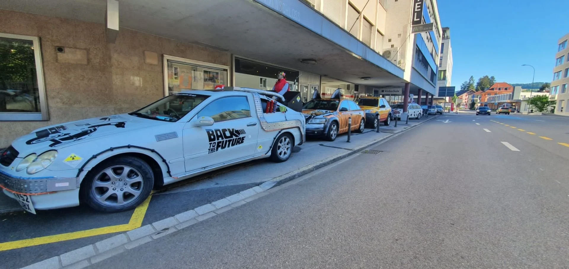A line of parked cars on a city street replacing their original appearances with various themed designs, including one resembling a 'Back to the Future' DeLorean.