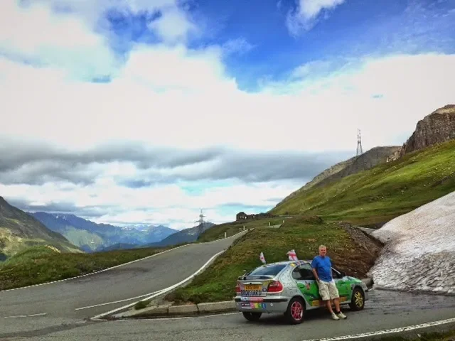 A man leaning on a silver hatchback car parked on a mountain roadside with a green landscape and mountains in the background, under a cloudy sky.