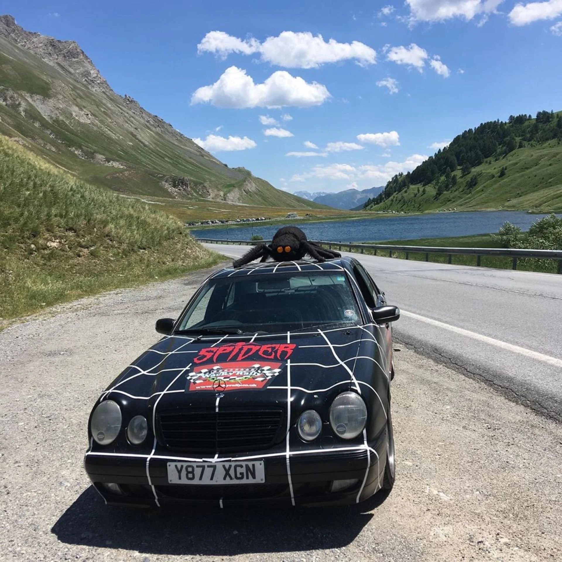 A black Mercedes-Benz car with a Spider-Man racing design parked beside a scenic mountain lake. A large Spider-Man figure with orange eyes is on the car's roof.