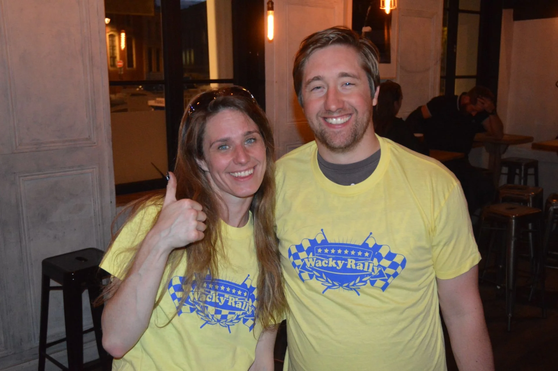 A smiling woman giving a thumbs-up and a smiling man stand close together inside a restaurant or cafe, both wearing yellow t-shirts with a blue graphic and the text 'Wacky Rally'.
