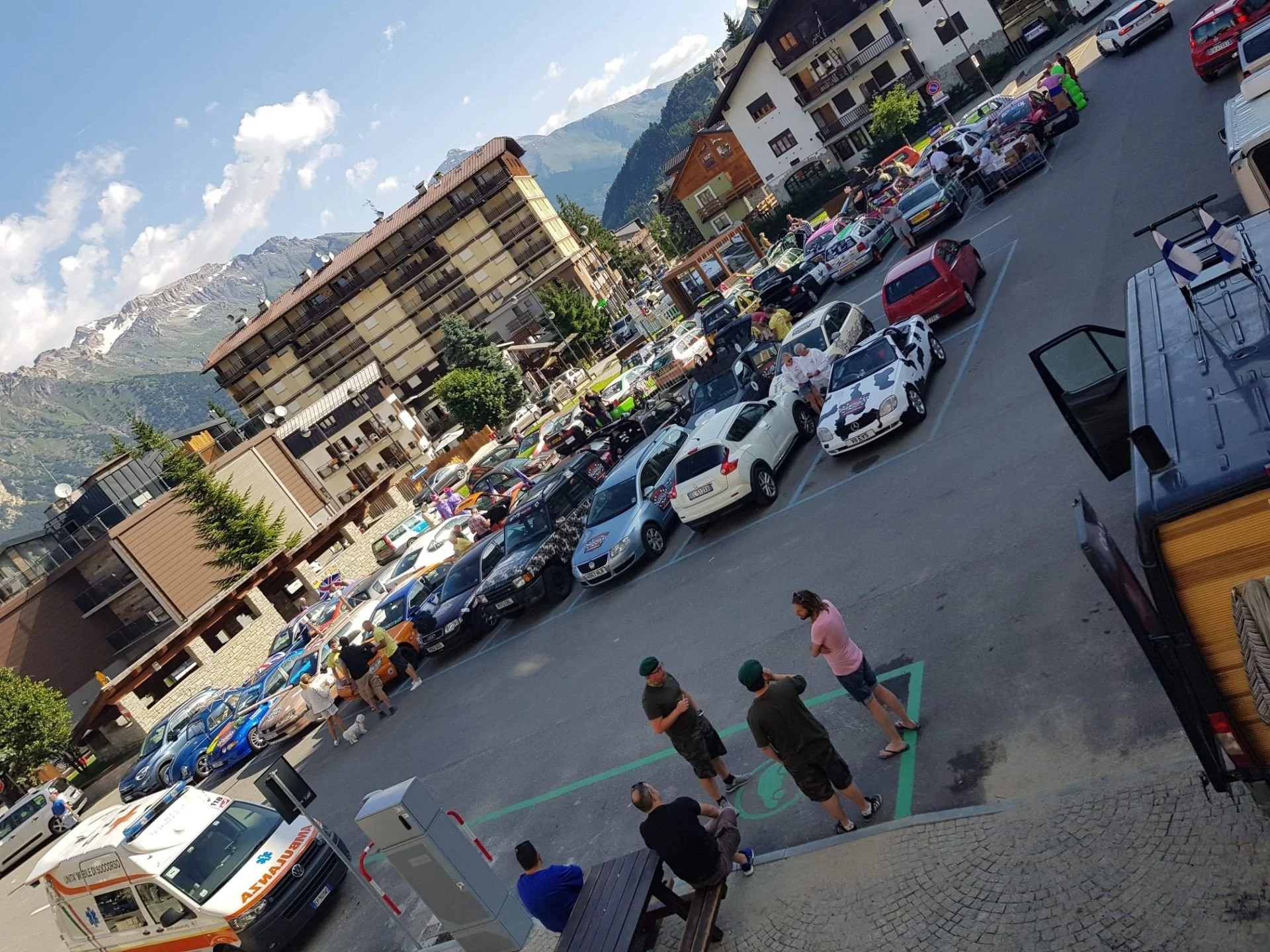 A parking lot filled with various cars, with several people standing and sitting, including two men in green caps talking to a woman in a pink shirt. In the background, there are buildings and mountains under a partly cloudy sky.
