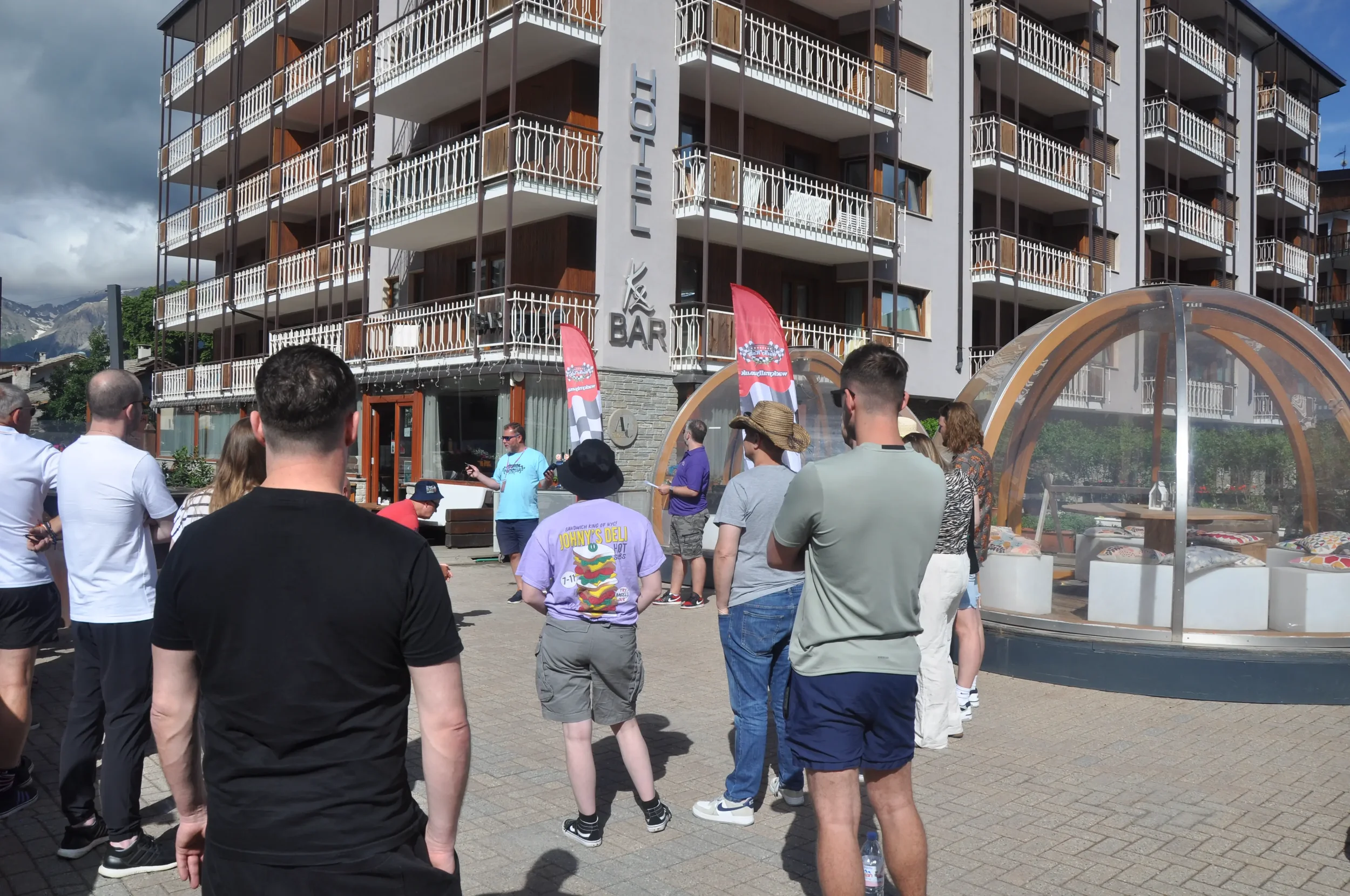 Group of people gathered outdoors in front of a building with a sign that says 'Hotel L' and 'Bar'. There is a man speaking to the crowd, and the building has multiple balconies and a mountain backdrop. Two pink flags are visible near the speaker, an
