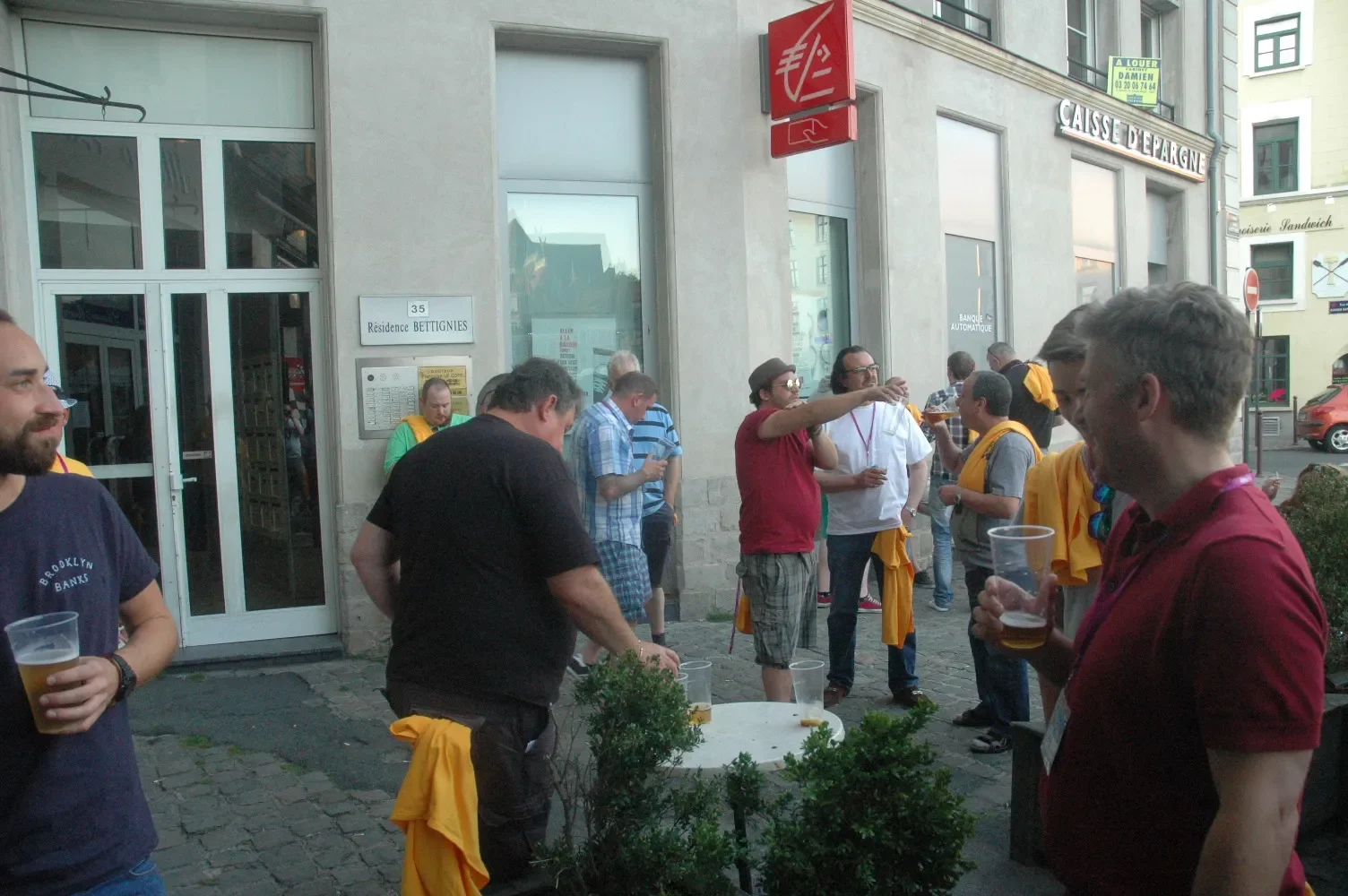 Group of people gathered outside a building, some holding plastic cups of beer, engaging in conversation during an outdoor event.