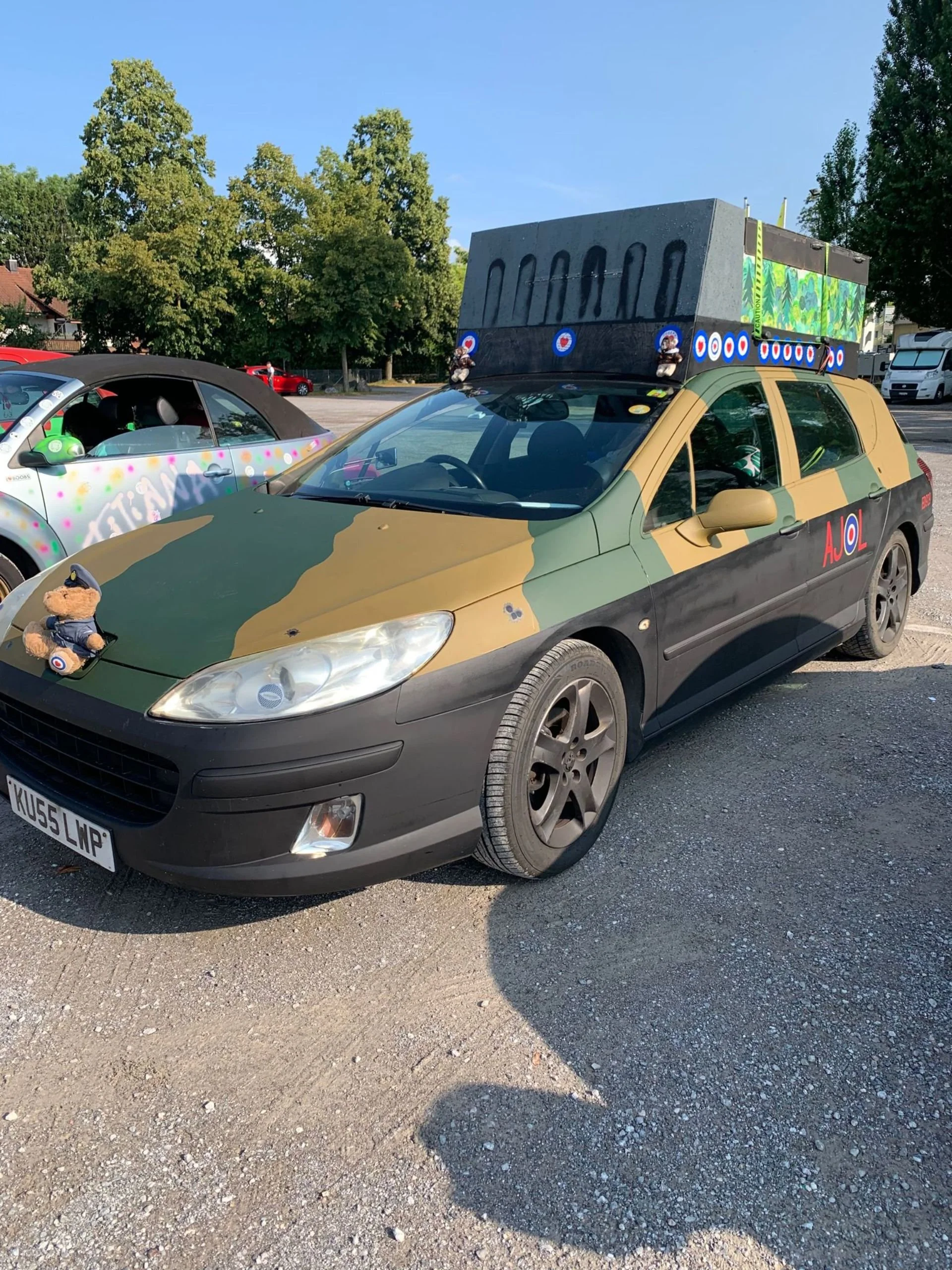 A car painted in camouflage colors with various military-themed decorations, including a large cardboard tank on the roof, a teddy bear with a military hat on the hood, and military emblem decals. The car is parked on a gravel lot with trees and othe