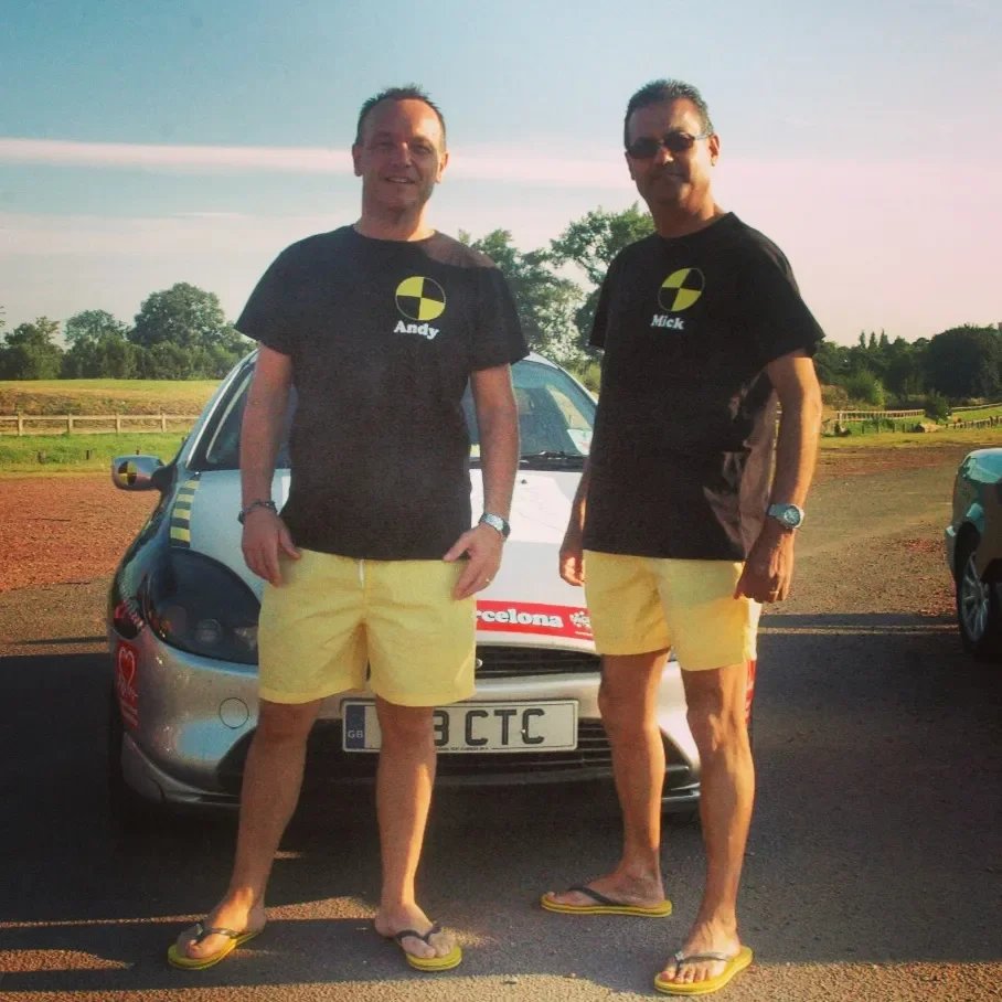 Two men wearing black t-shirts and yellow shorts standing in front of a car outdoors during daytime.