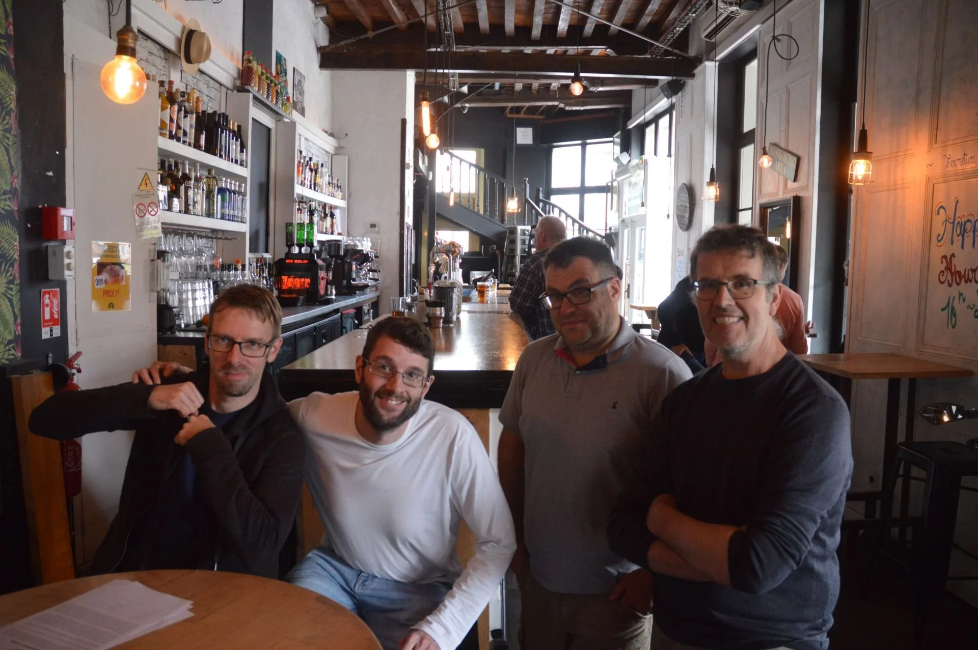 Four men sitting at a table in a cozy cafe bar, with a counter and barista equipment behind them, in a rustic, warmly lit space with large windows.