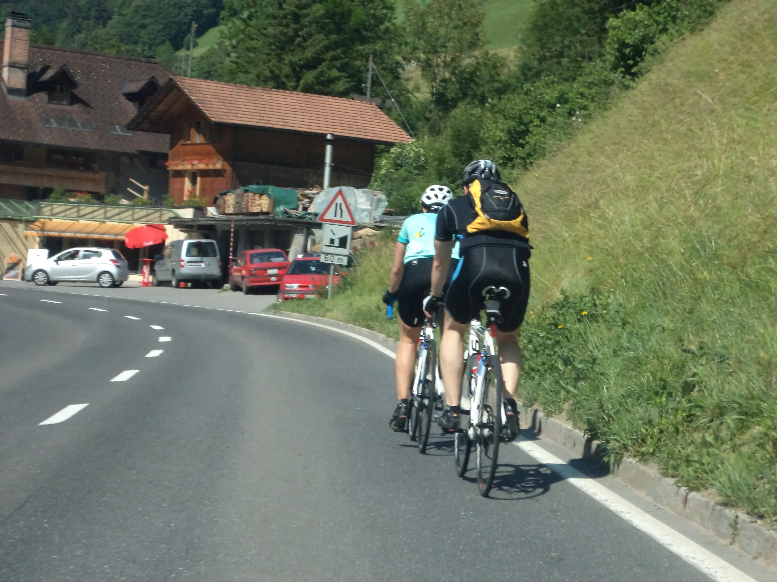 Two cyclists riding on a curved mountain road with houses and green hills in the background.