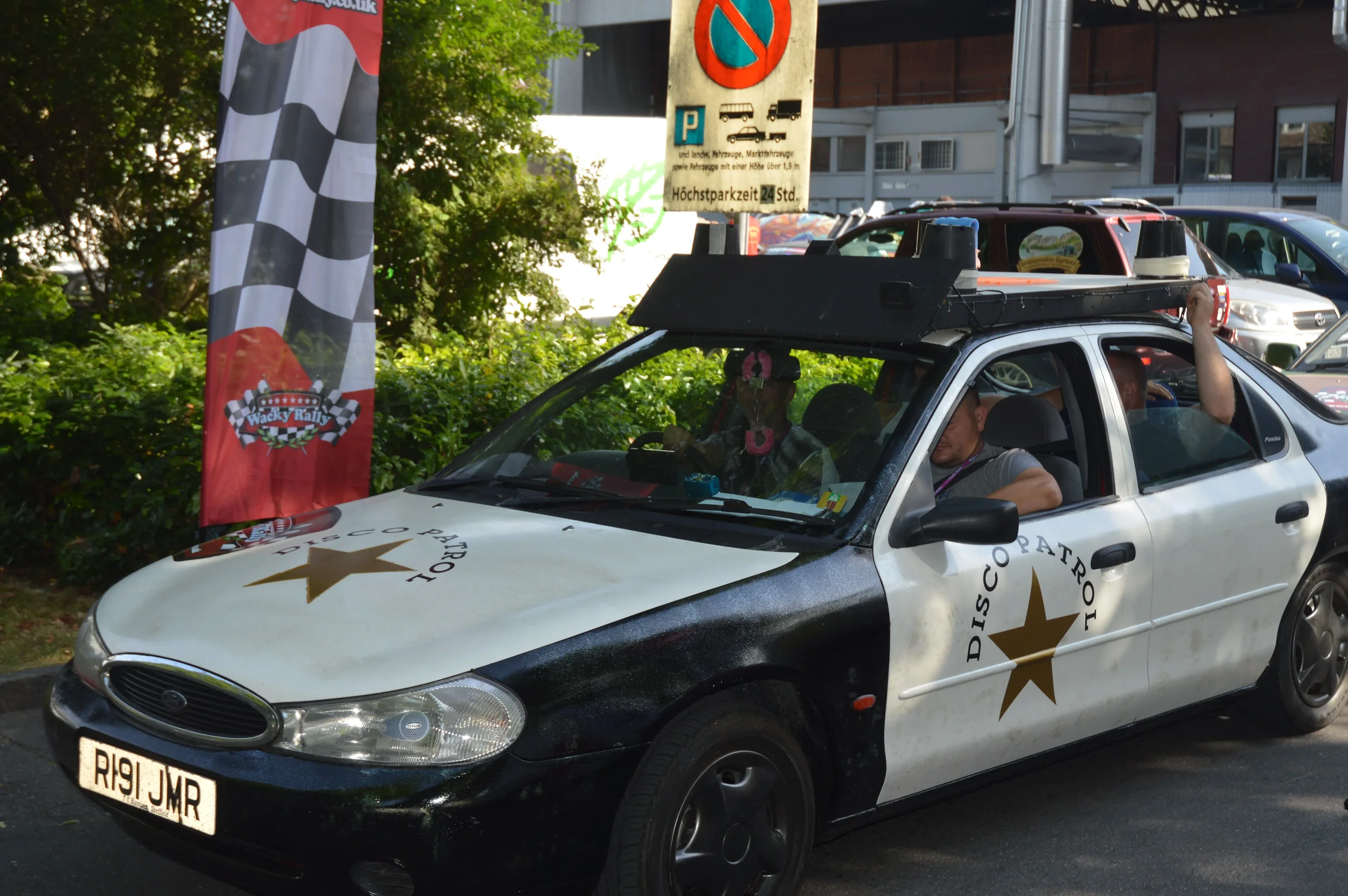 A modified police car with a black and white color scheme and large 'DISCO PATROL' logos on the doors, parked on a street with a red and black checkered flag banner and parking signs in the background.