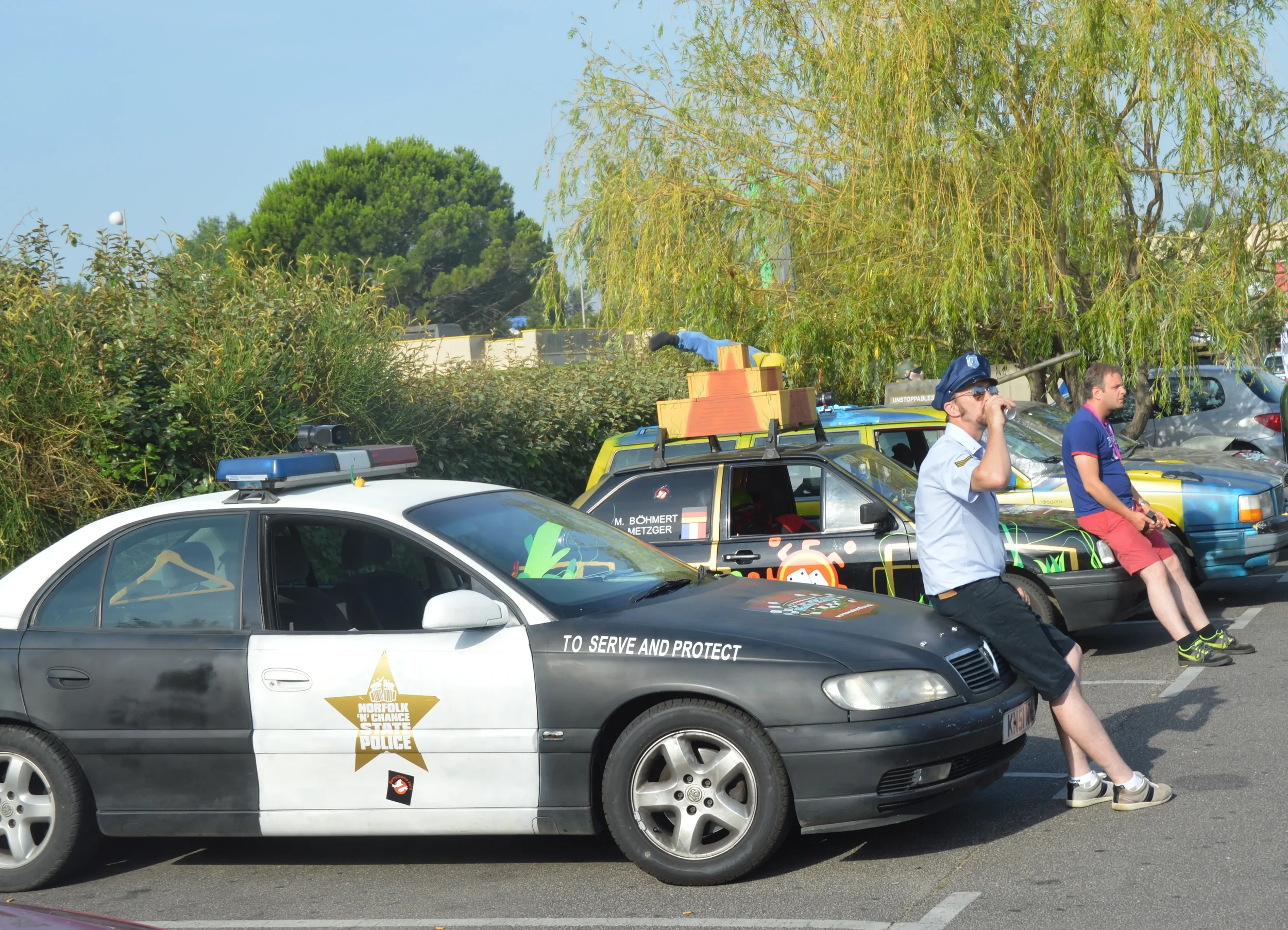 A variety of decorated police cars parked outside, with two young men sitting on the cars, one drinking and the other relaxing, in a parking lot with trees in the background.