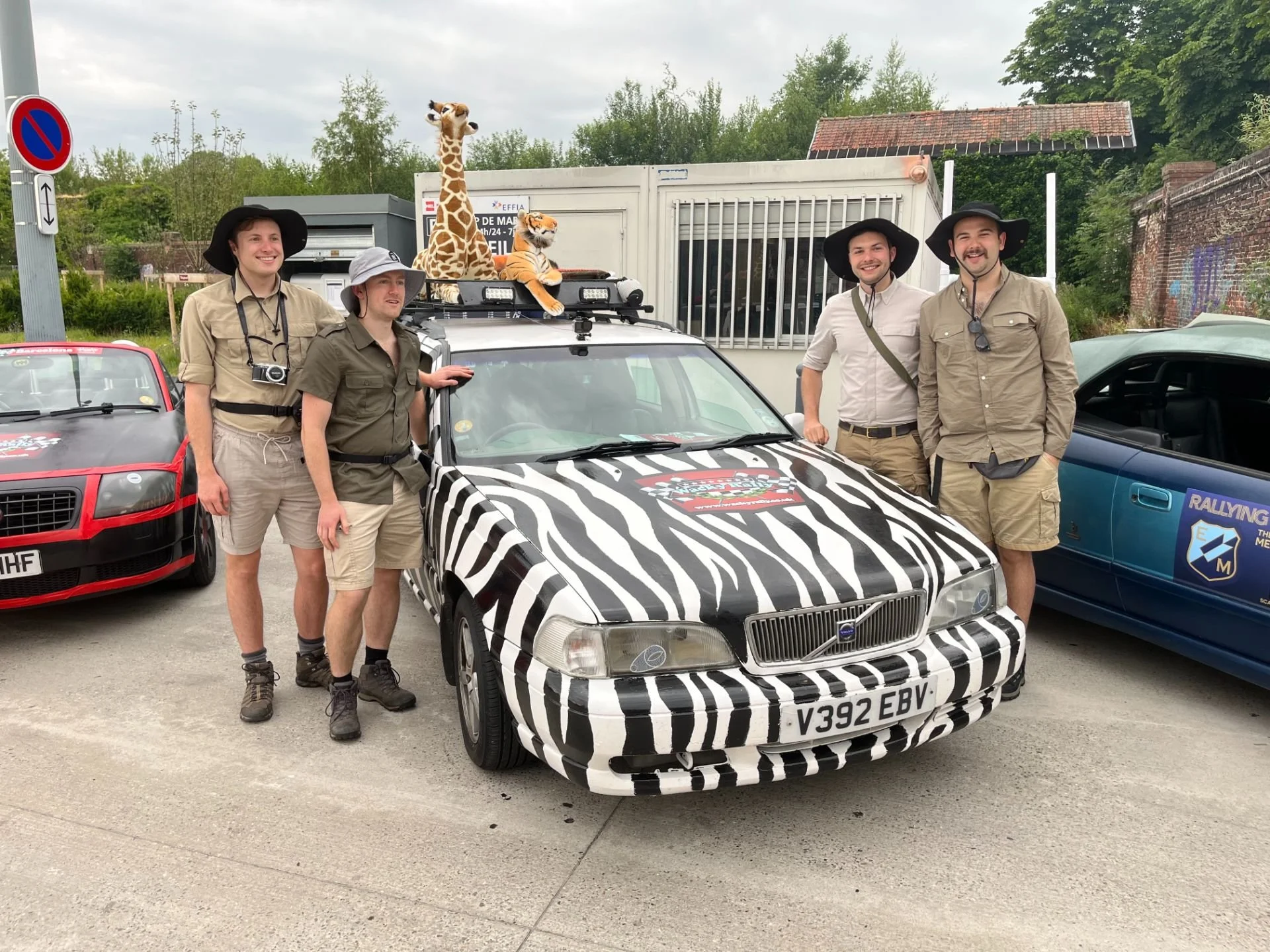Group of five men in safari outfits standing next to a zebra-striped rally car with plush giraffe and tiger on top, parked among other cars, outdoors under cloudy sky.