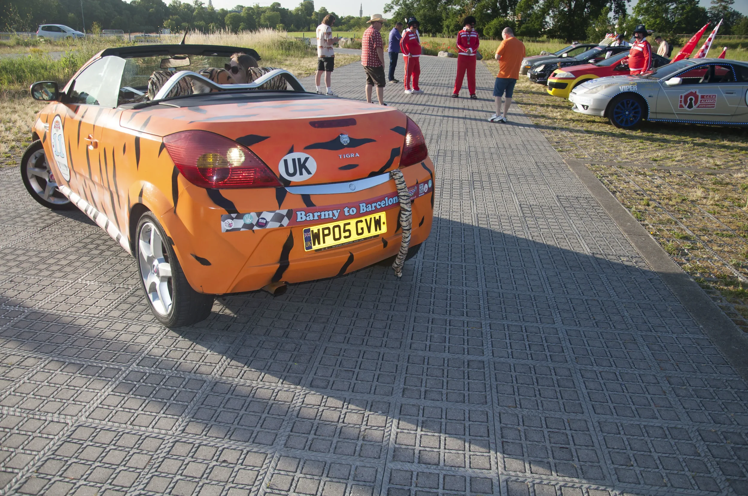 A parking lot filled with small, uniquely decorated cars, with a focus on an orange convertible with tiger stripe paint, a bumper sticker reading 'Barmy to Barcelona,' and license plate 'WP05 GVW.' Several people are gathered around, engaging and obs