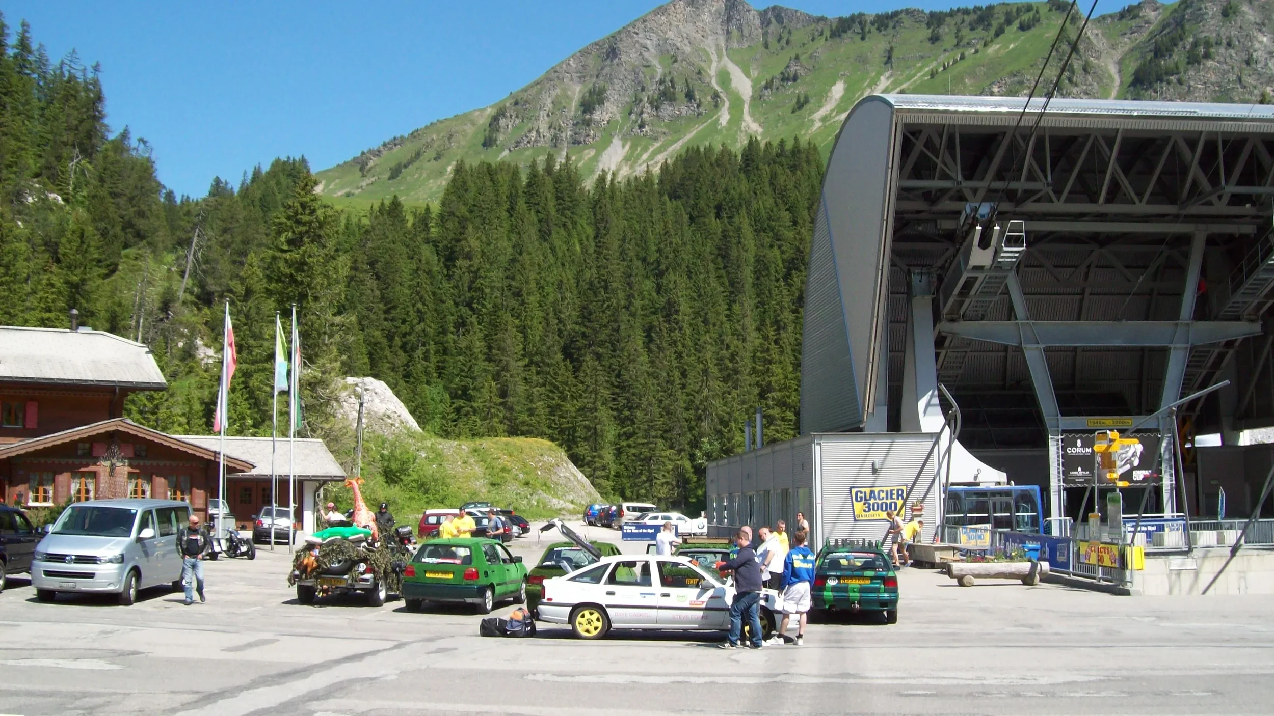 Parking lot at a mountain ski resort with cars and people near a ski lift, surrounded by green trees and mountain scenery.
