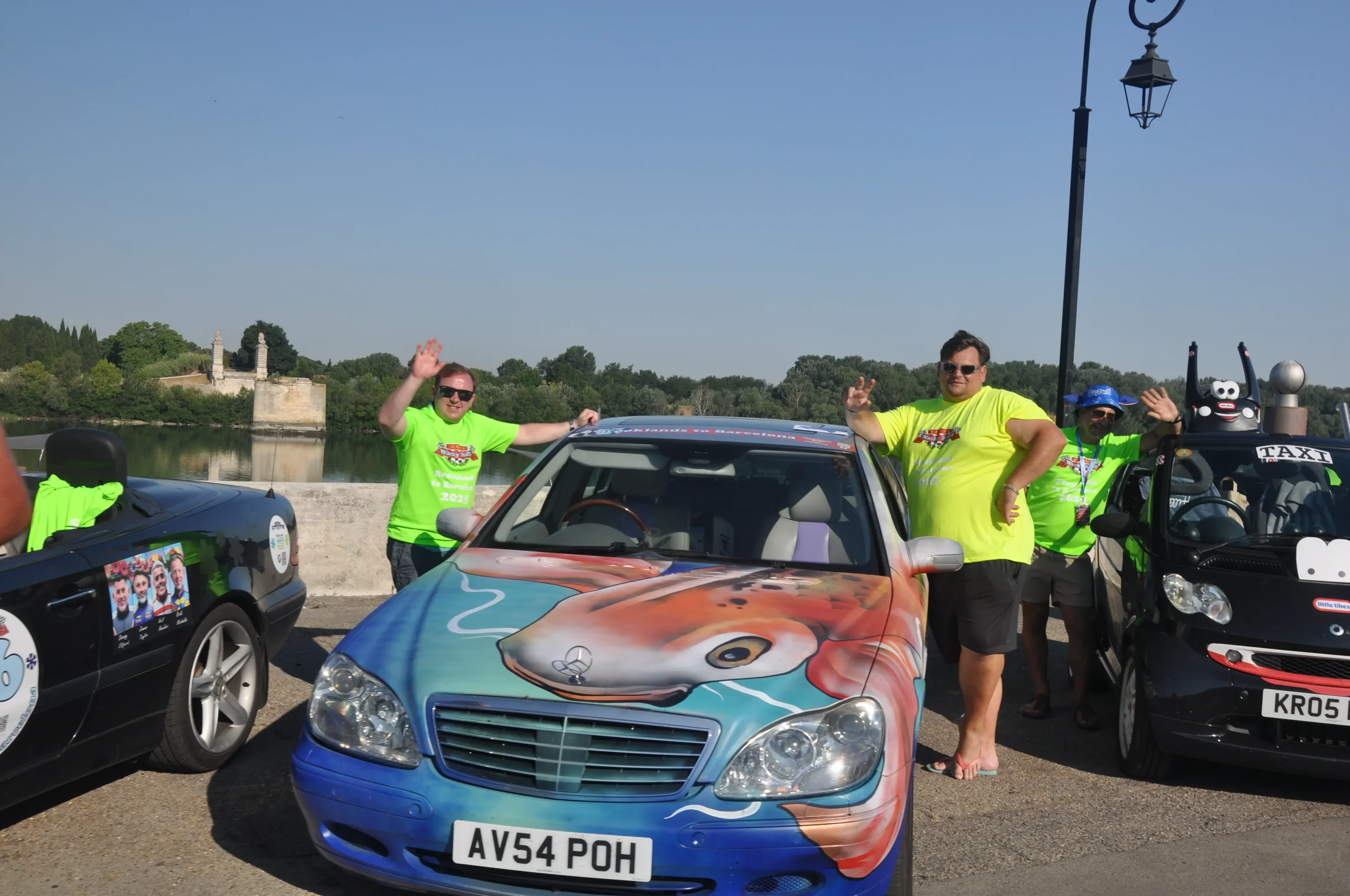 Three people standing near colorful decorated cars, waving at the camera during a sunny outdoor event, with trees and a river in the background.