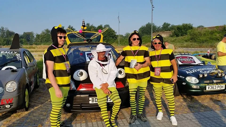 Four people dressed in coordinated bee-themed costumes, standing in front of a decorated car at an outdoor event.