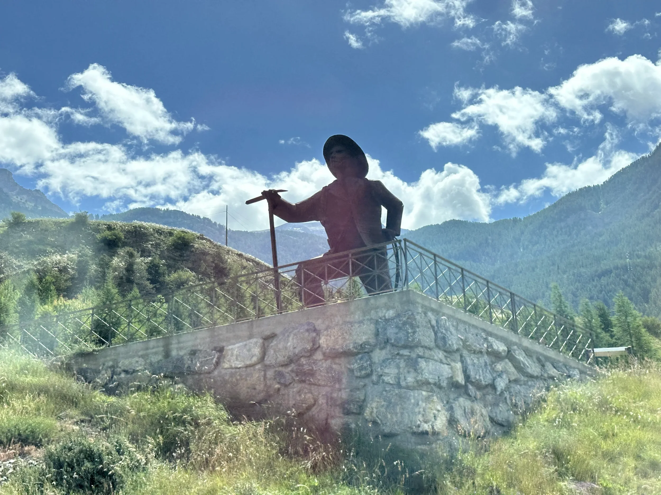 A large bronze statue of a person wearing a hat, holding a long stick, located on a stone platform with a metal railing, set against a mountainous landscape with blue sky and white clouds.