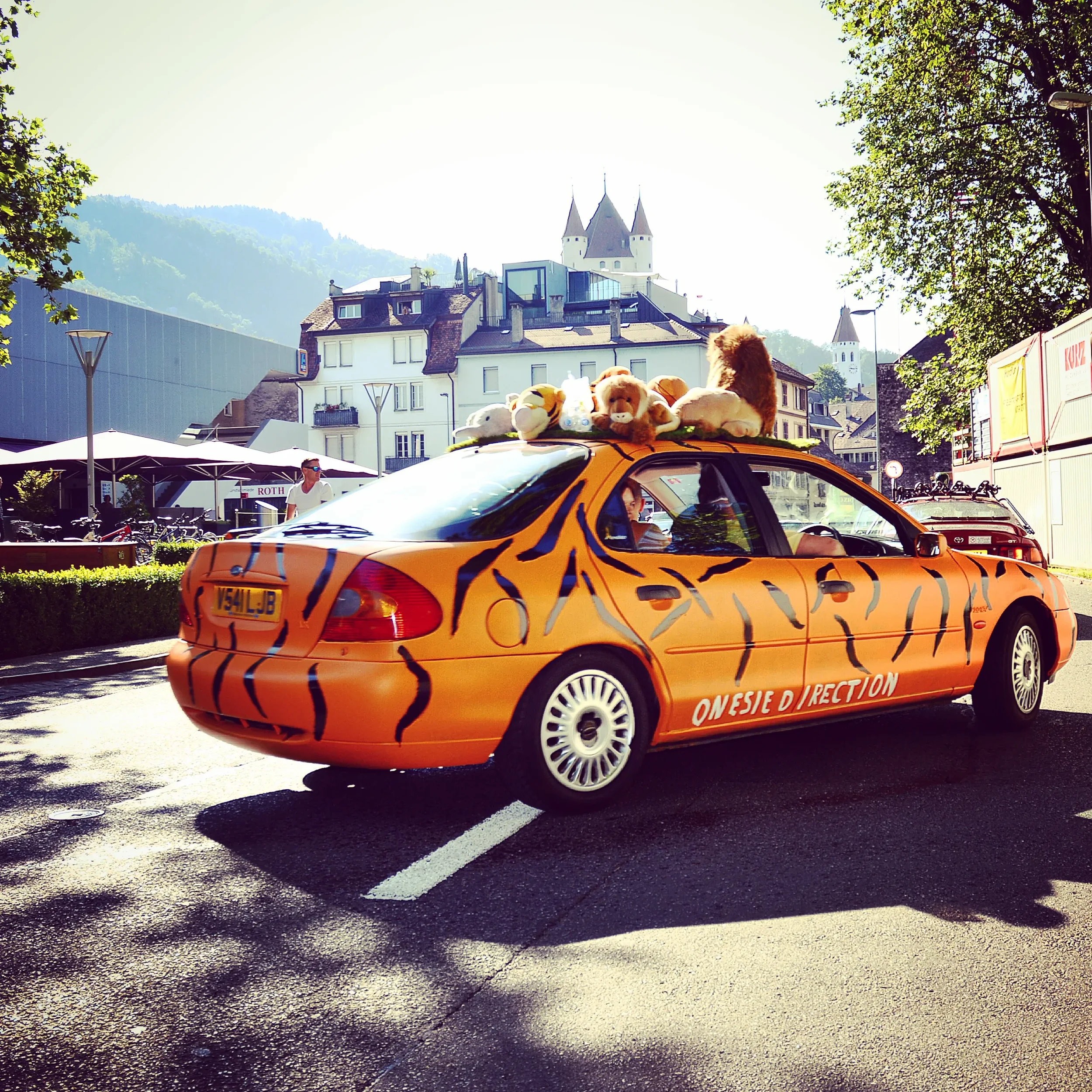 Orange car with black tiger stripe design, decorated with plush toys on roof, parked on street with buildings and trees in background.