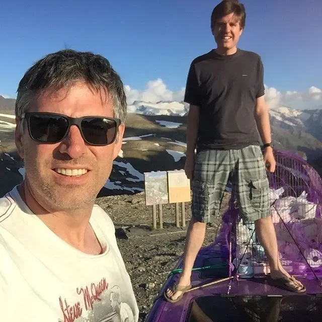Two men smiling at the camera with a mountainous landscape in the background, one standing on a purple car and the other taking the selfie.