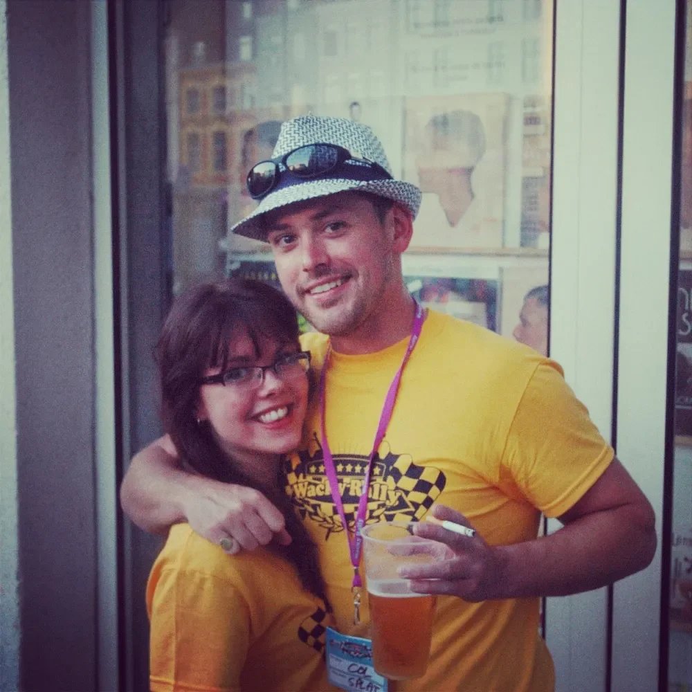 A man and woman smiling and hugging at an outdoor event, both wearing yellow shirts, the man with sunglasses on his hat, holding a beer, and the woman wearing glasses.